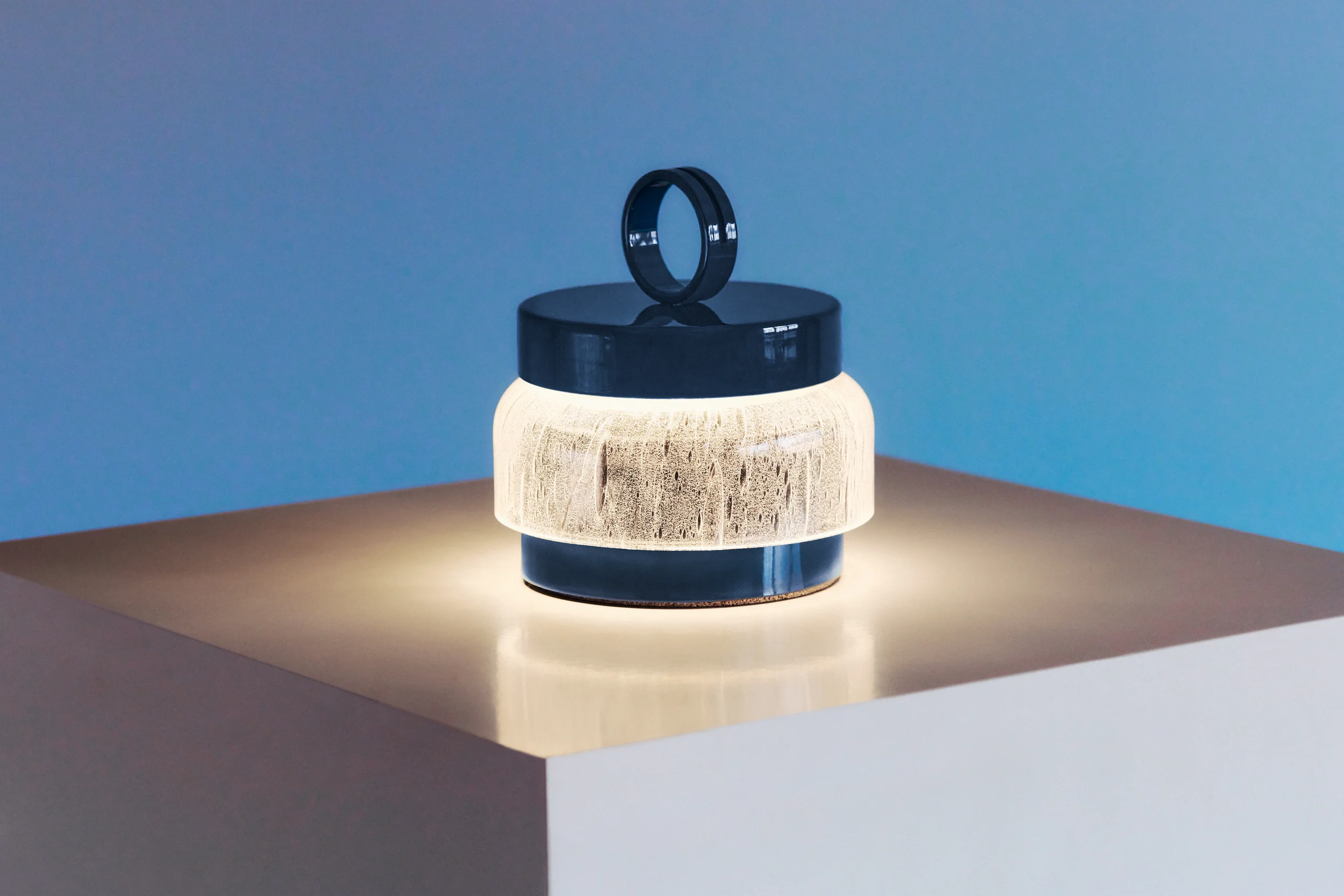 A modern table lamp with a blue handle on top, a textured white middle section, and a dark base, placed on a beige surface against a blue background.