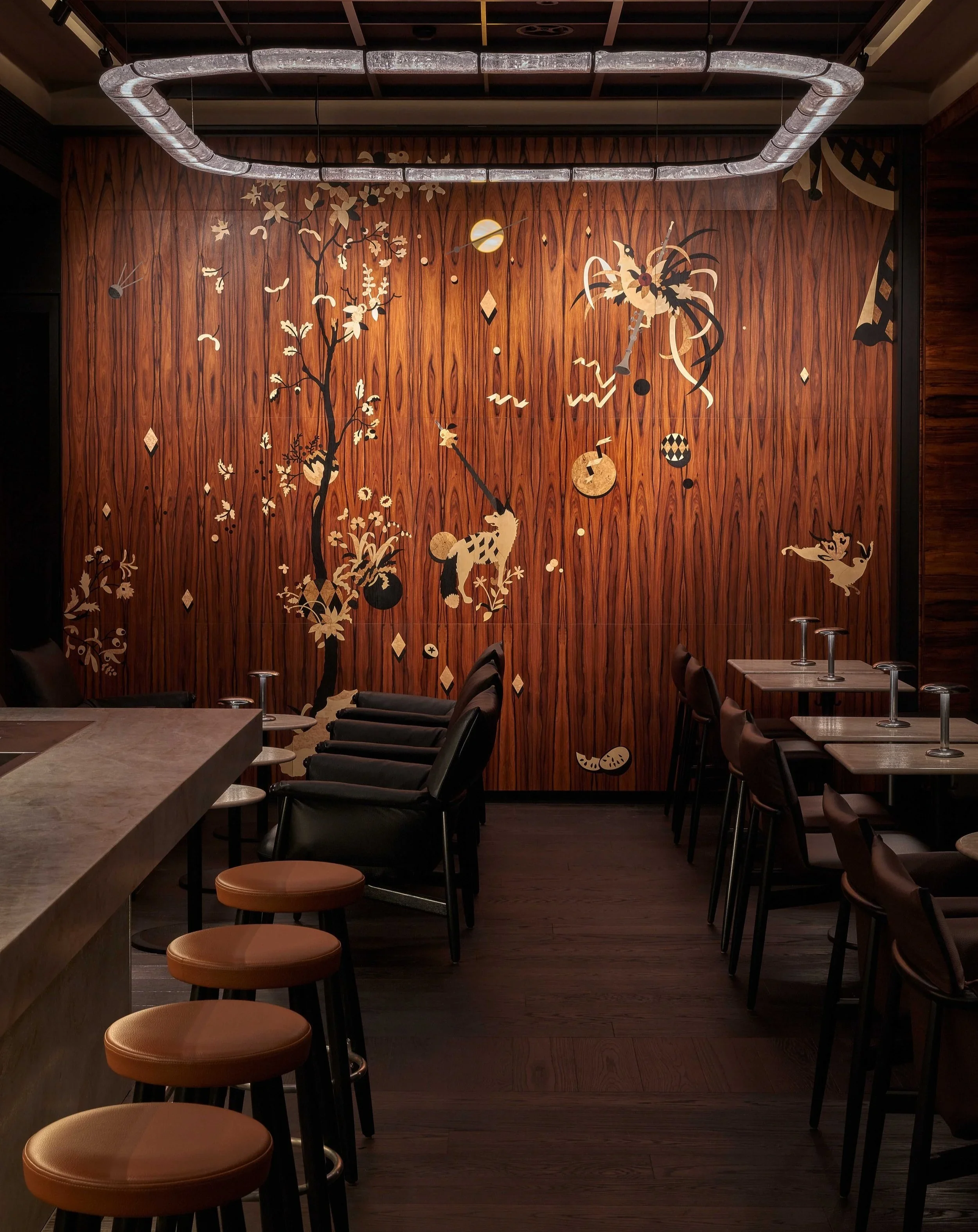 Interior of a restaurant with a wooden wall mural featuring Japanese-style art of trees, animals, and geometric patterns. There are black chairs and brown stools around tables.