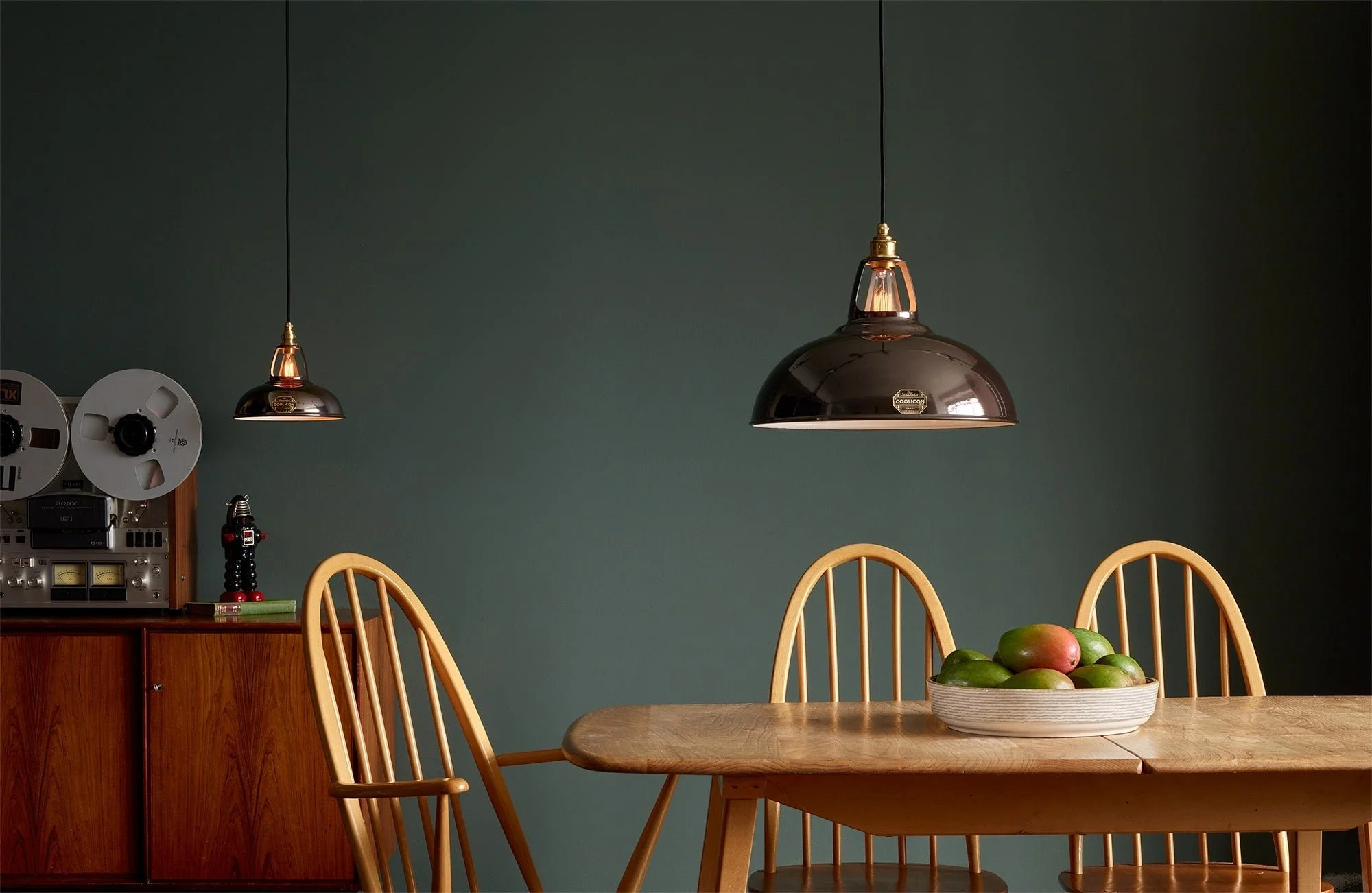 A wooden dining table with six matching chairs, a ceramic bowl filled with mangoes, and a wooden sideboard with vintage audio equipment and a nutcracker, green wall in the background.