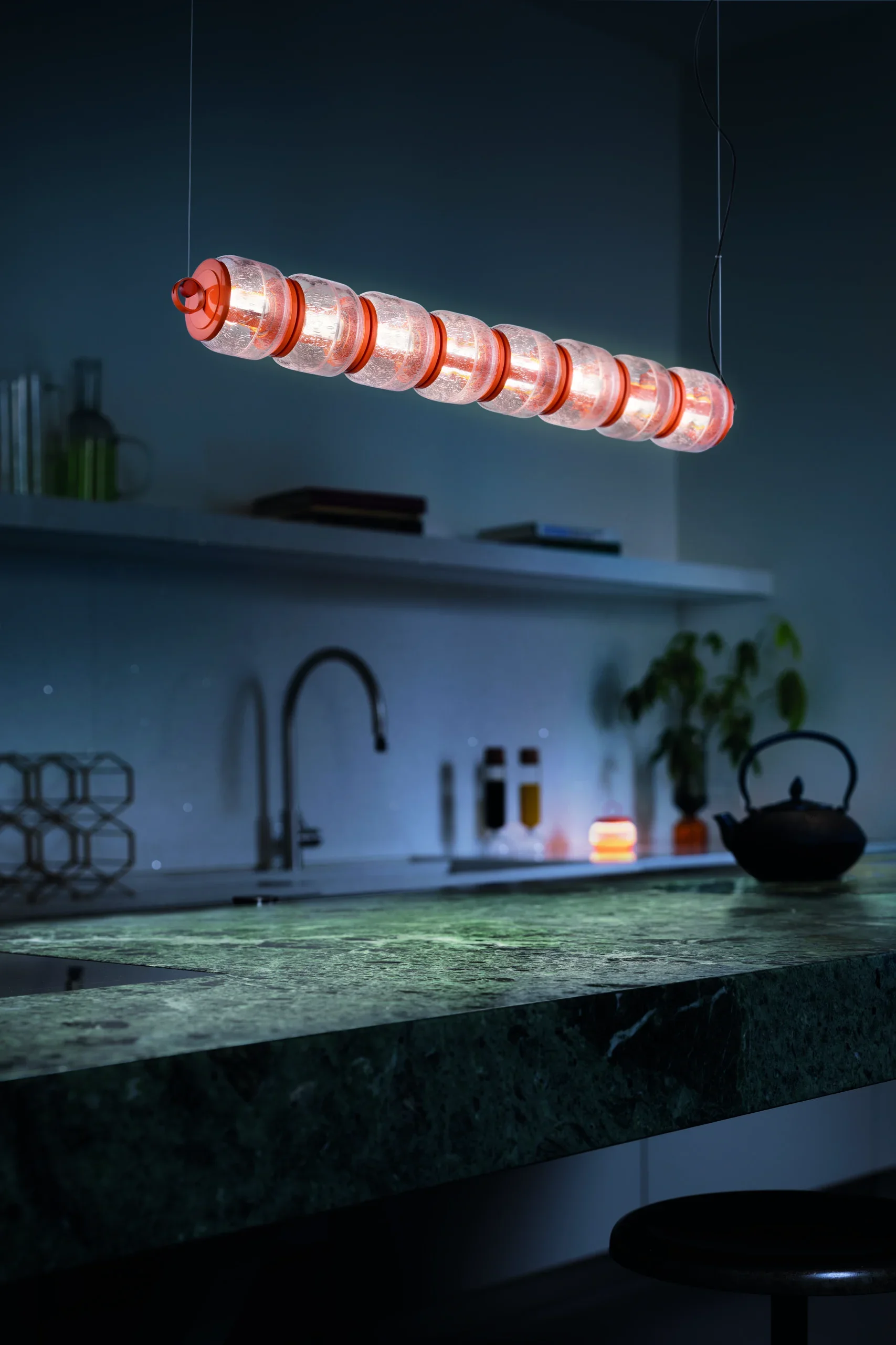 A modern kitchen with a hanging light fixture made of glass and metal, a dark green marble countertop, a black teapot, and some kitchen utensils and plants in the background.