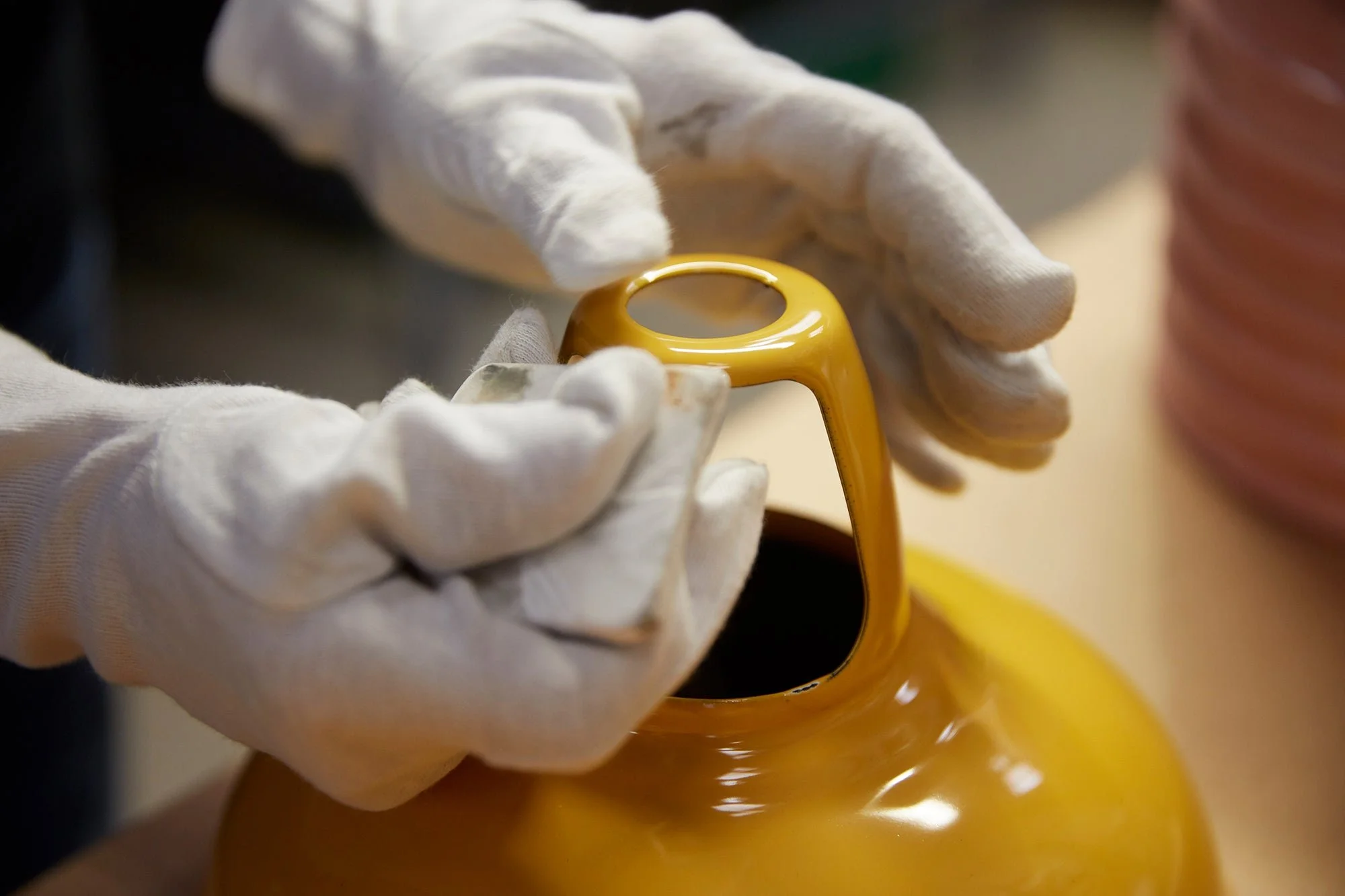 Person wearing white gloves polishing a yellow enamel lamp shade.