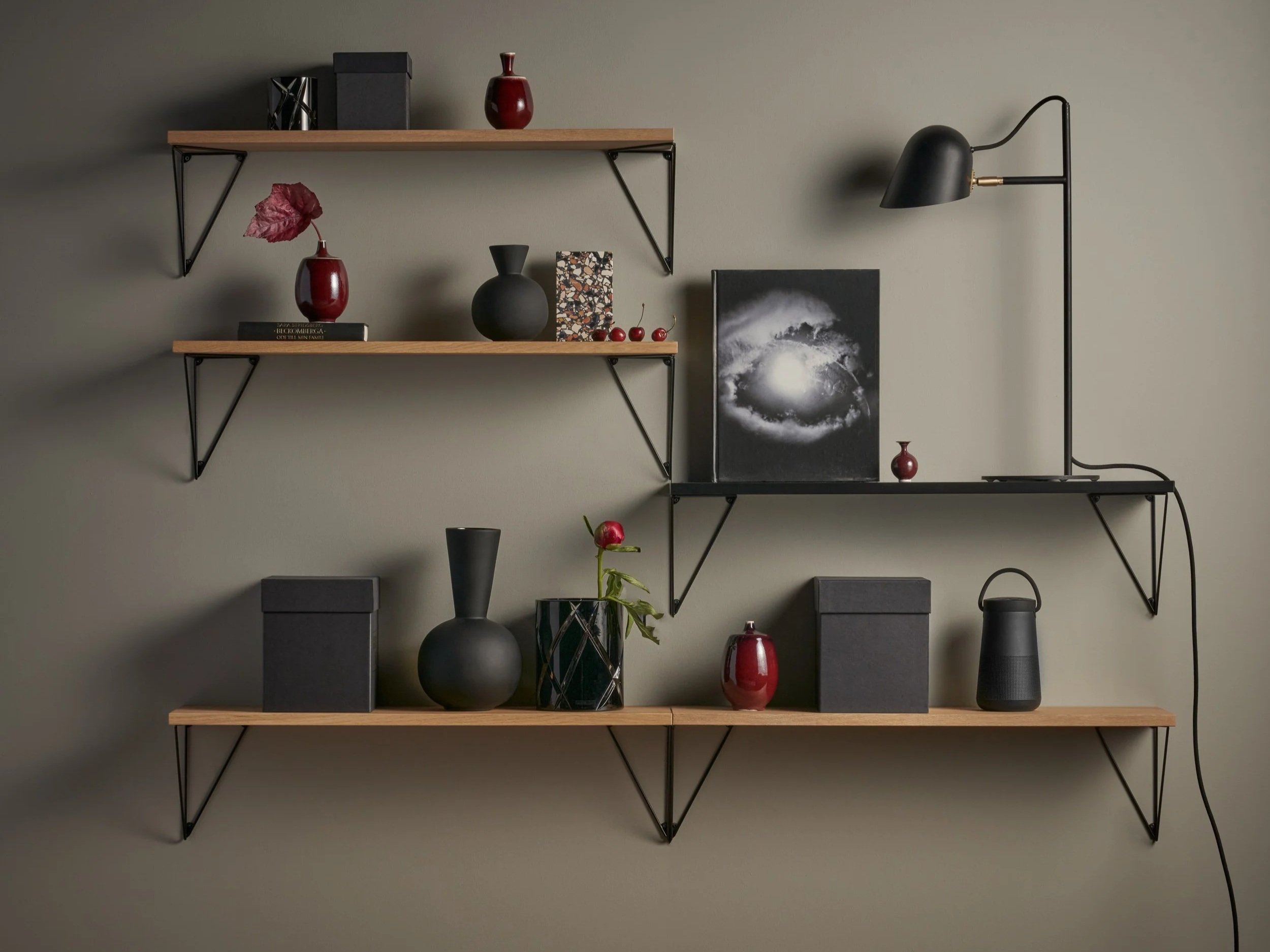 Two wooden wall-mounted shelves with black metal brackets hold decorative vases and artwork on a muted gray wall. A black wall-mounted lamp with a flexible arm is positioned on the right.