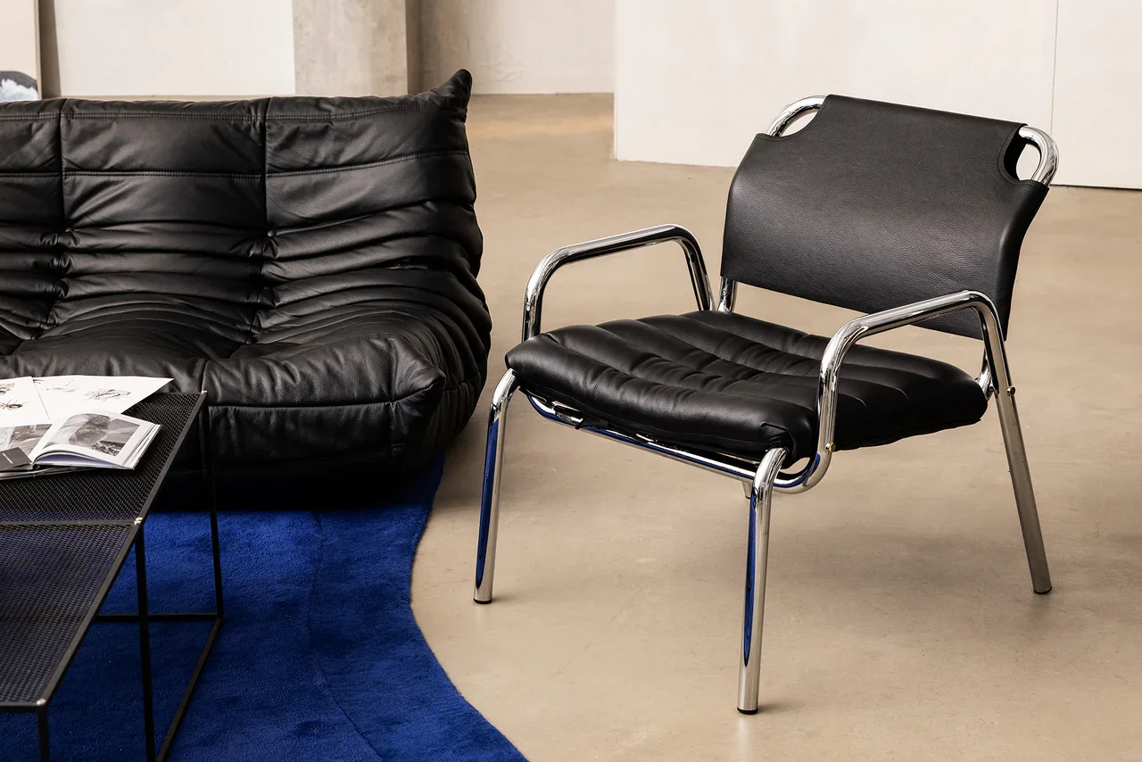Modern black leather armchair and black office chair with chrome legs in a room with a gray floor, white walls, blue rug, and a small black table with magazines.