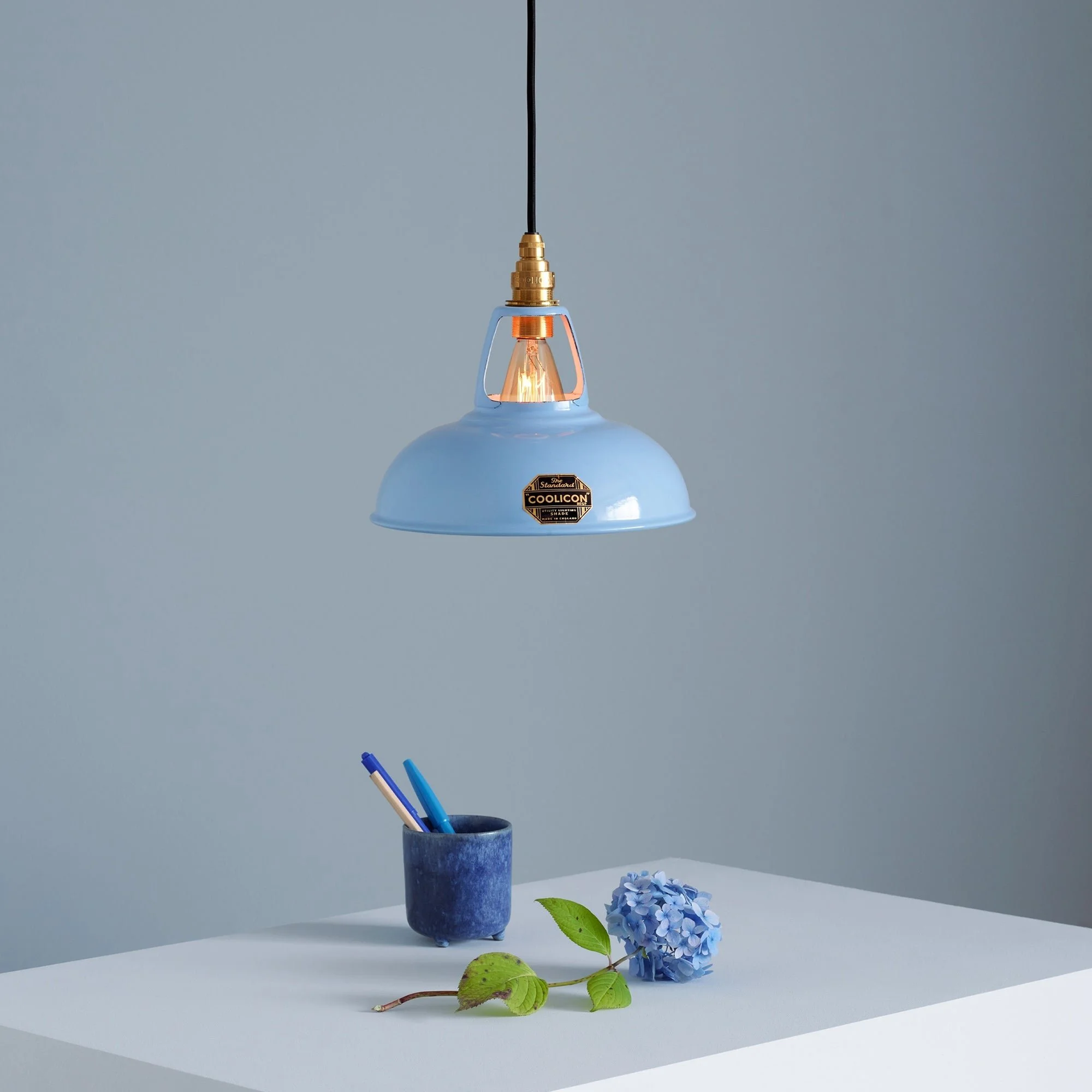 A blue pendant lamp hanging above a white table. On the table, there is a blue ceramic cup holding two pens, and a blue hydrangea flower with green leaves placed on the surface.
