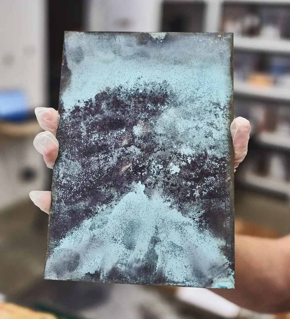 Person holding a rectangular metal object with abstract black and blue paint splatter pattern, in a laboratory or workshop setting.