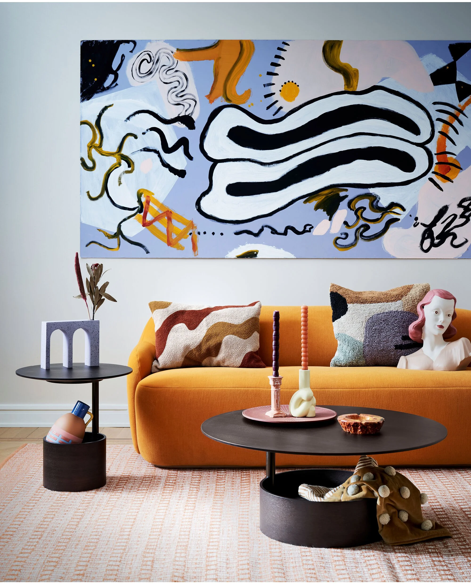 Modern living room with an orange sofa, abstract artwork on the wall, colorful throw pillows, a black side table with decor, a round black coffee table with candles and a decorative object, and a textured rug.