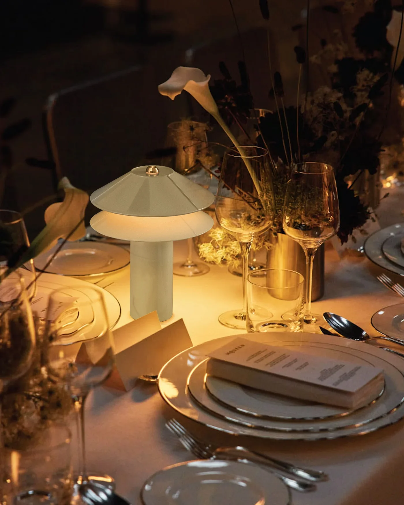 A dinner table set with plates, cutlery, glasses, and a small modern lamp, illuminated by soft warm light with flowers and a menu on the table.