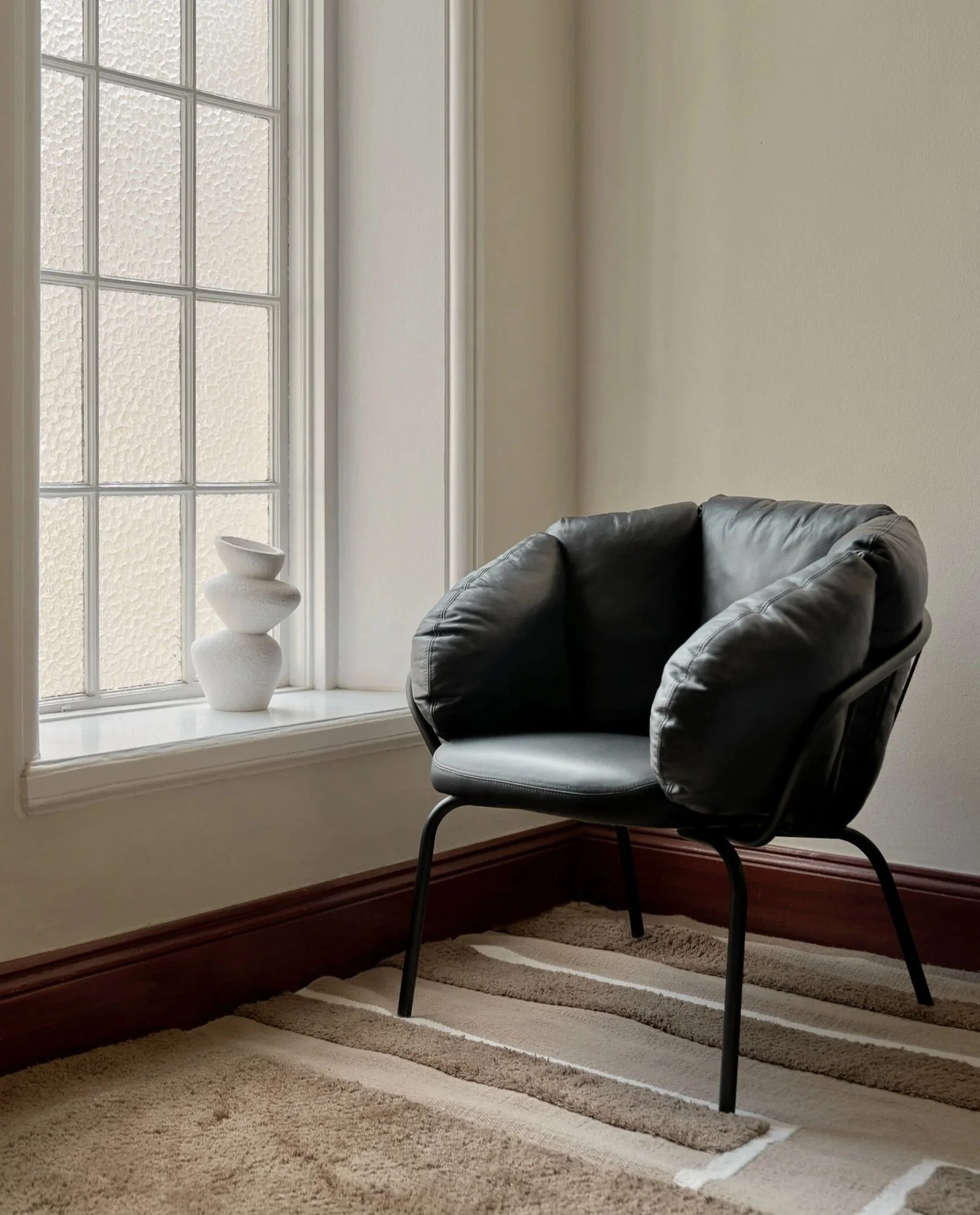 A black lounge chair next to a window with textured glass and a white decorative sculpture on the windowsill, in a room with beige walls and a striped beige and brown carpet.