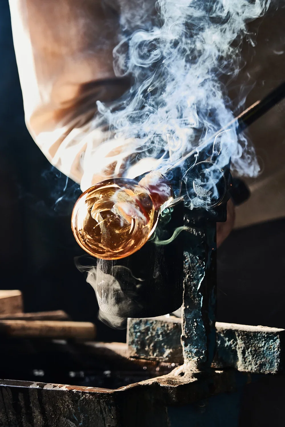 Close-up of a blacksmith forging with a glowing glass object emitting smoke, with sparks and heat visible.