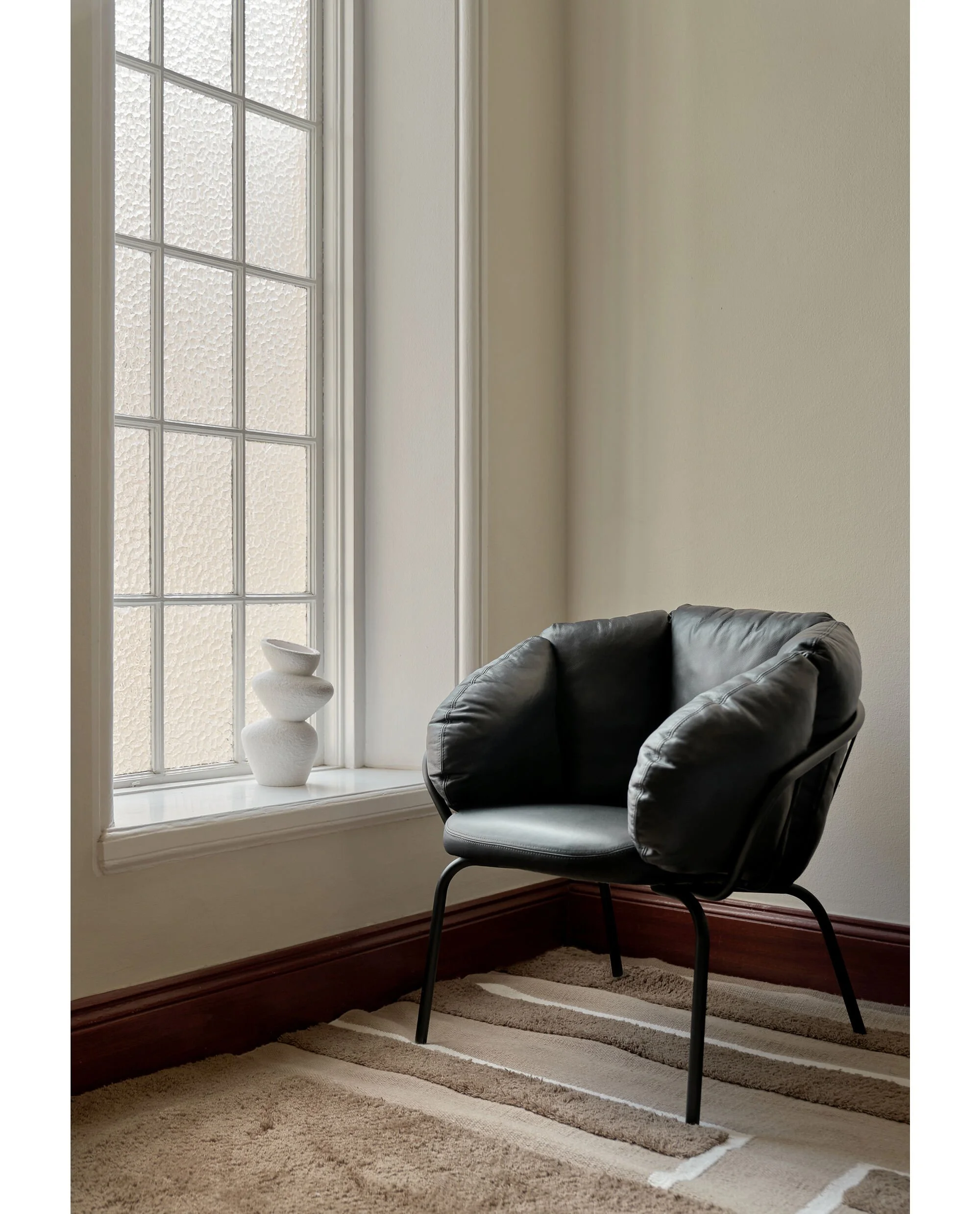 A black modern armchair next to a window with textured glass, with a white ceramic sculpture on the windowsill, beige walls, and a patterned beige and brown rug.