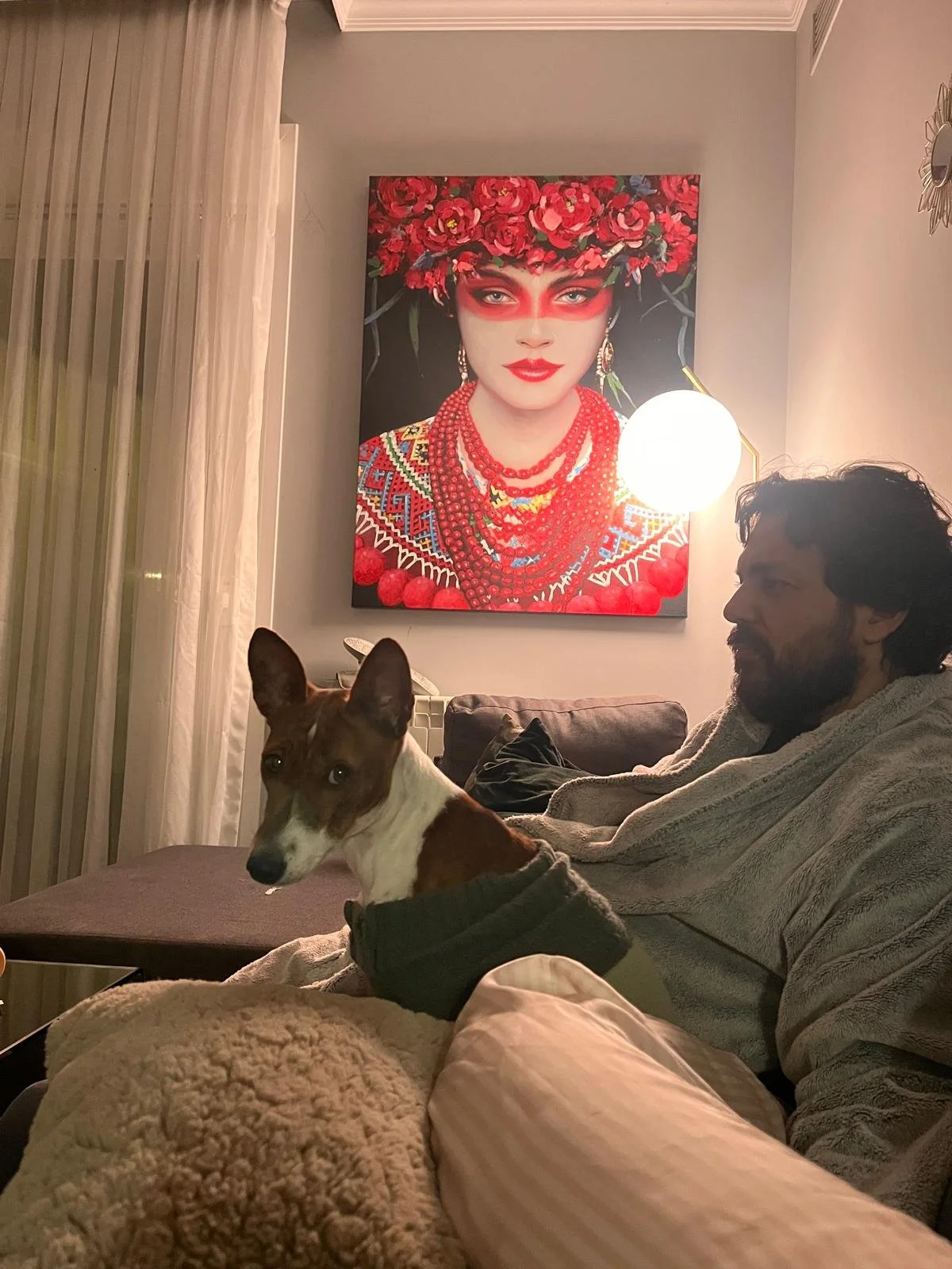 Un hombre con un perro en un sillón frente a una pintura de una mujer vestida con estilo tradicional y accesorios de color rojo