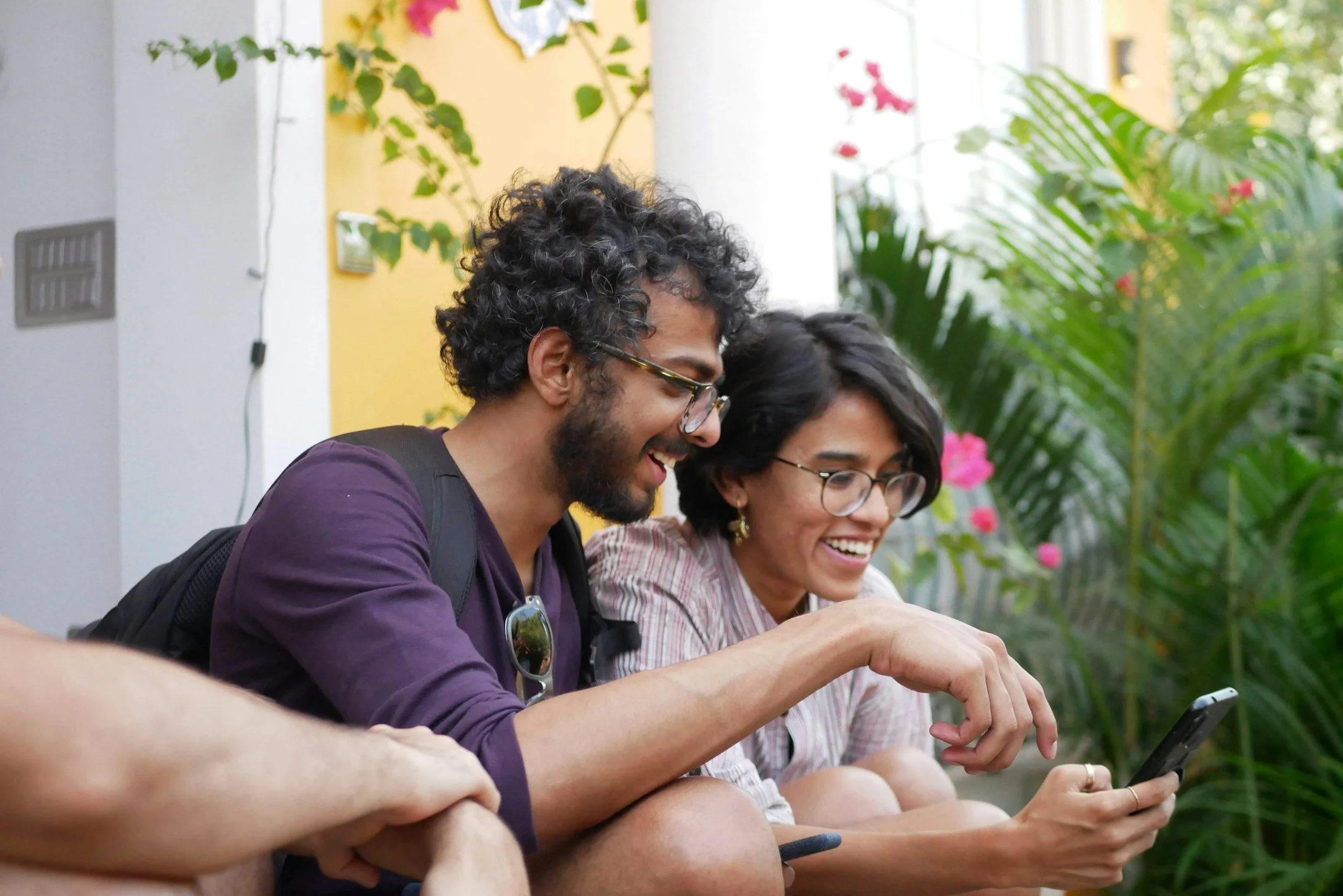 Two young adults, a man and a woman, sitting outdoors, smiling and looking at a smartphone together.