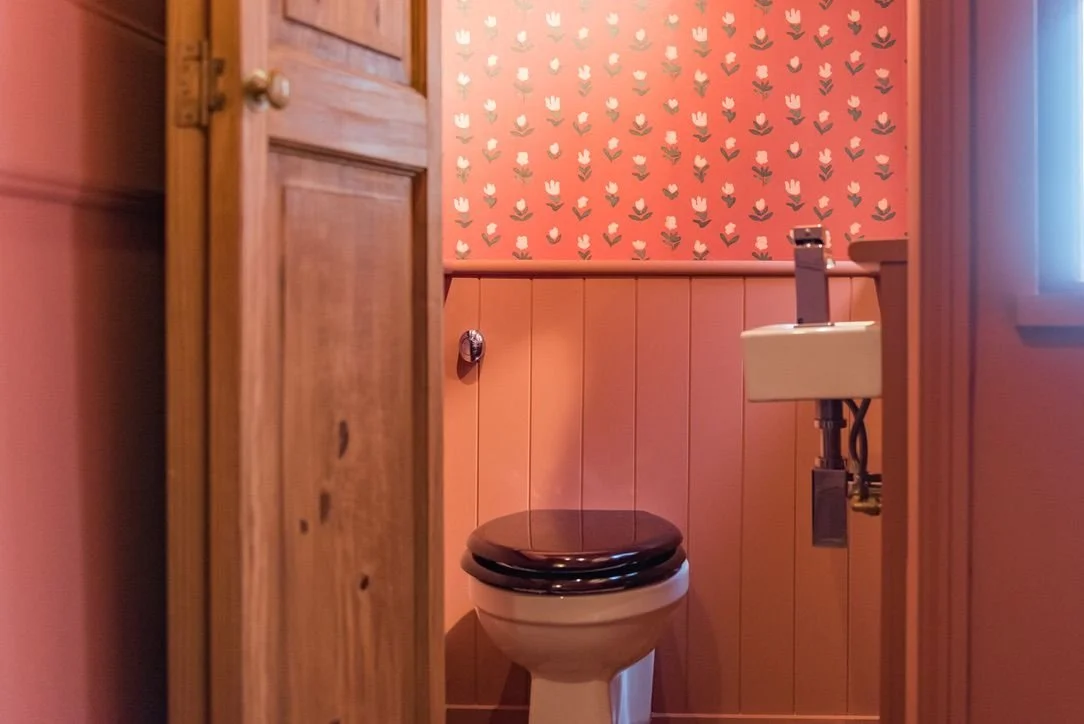 View of a small bathroom with a toilet and a small sink, decorated with pink and floral wallpaper and wood-paneled walls.