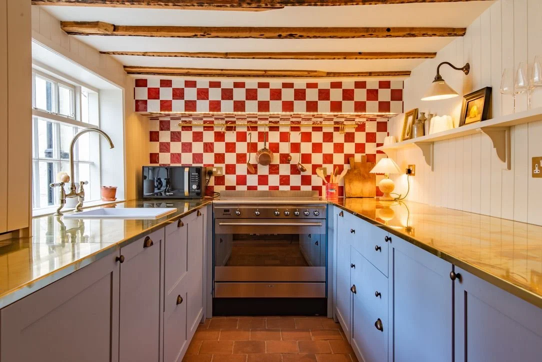 Cozy kitchen with white cabinets, wooden countertops, a checkered red and white backsplash, and exposed wooden ceiling beams.