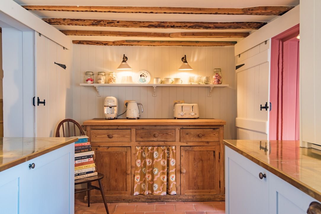 Rustic kitchen with white paneled walls, exposed wood ceiling beams, and a central wooden cabinet. The cabinet has a floral curtain and a countertop with appliances, including a toaster, a coffee maker, and an electric kettle. Above, a shelf with gla