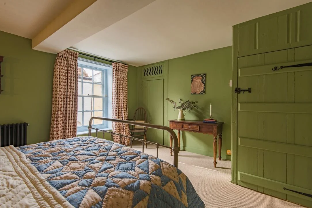 A bedroom with green walls, a large window with patterned curtains, a wooden headboard, a quilted bedspread, a wooden chair, a wooden console table with a vase of greenery, books, and a candle, and a small framed artwork on the wall.
