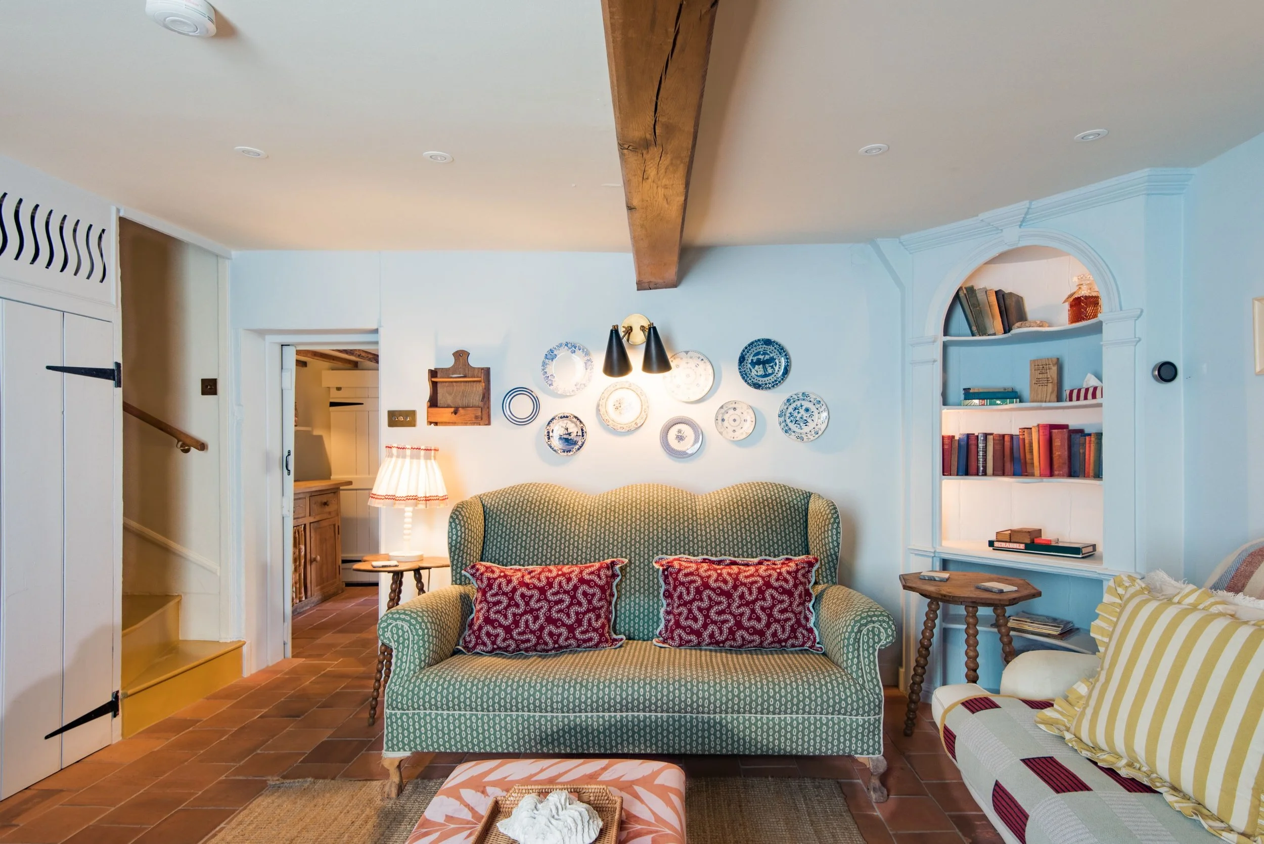 Living room with green patterned sofa, yellow and white striped armchair, wall-mounted plates, wooden ceiling beam, and built-in bookshelf.