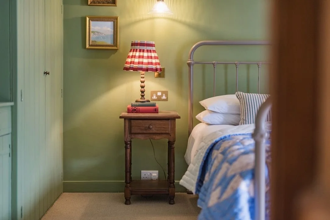 A cozy bedroom with green walls, a metal bed frame, a wooden nightstand, a red checkered lampshade, and framed paintings on the wall.