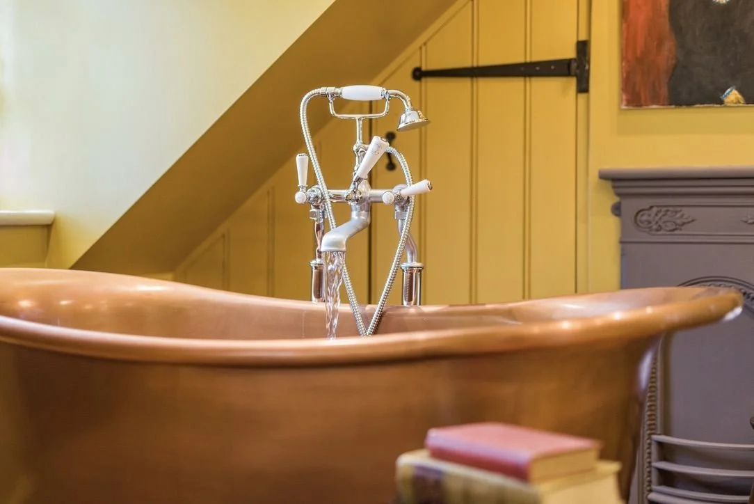 A copper bathtub with a vintage-style faucet and handheld showerhead, with a pink book nearby and beige paneling on the wall in the background.
