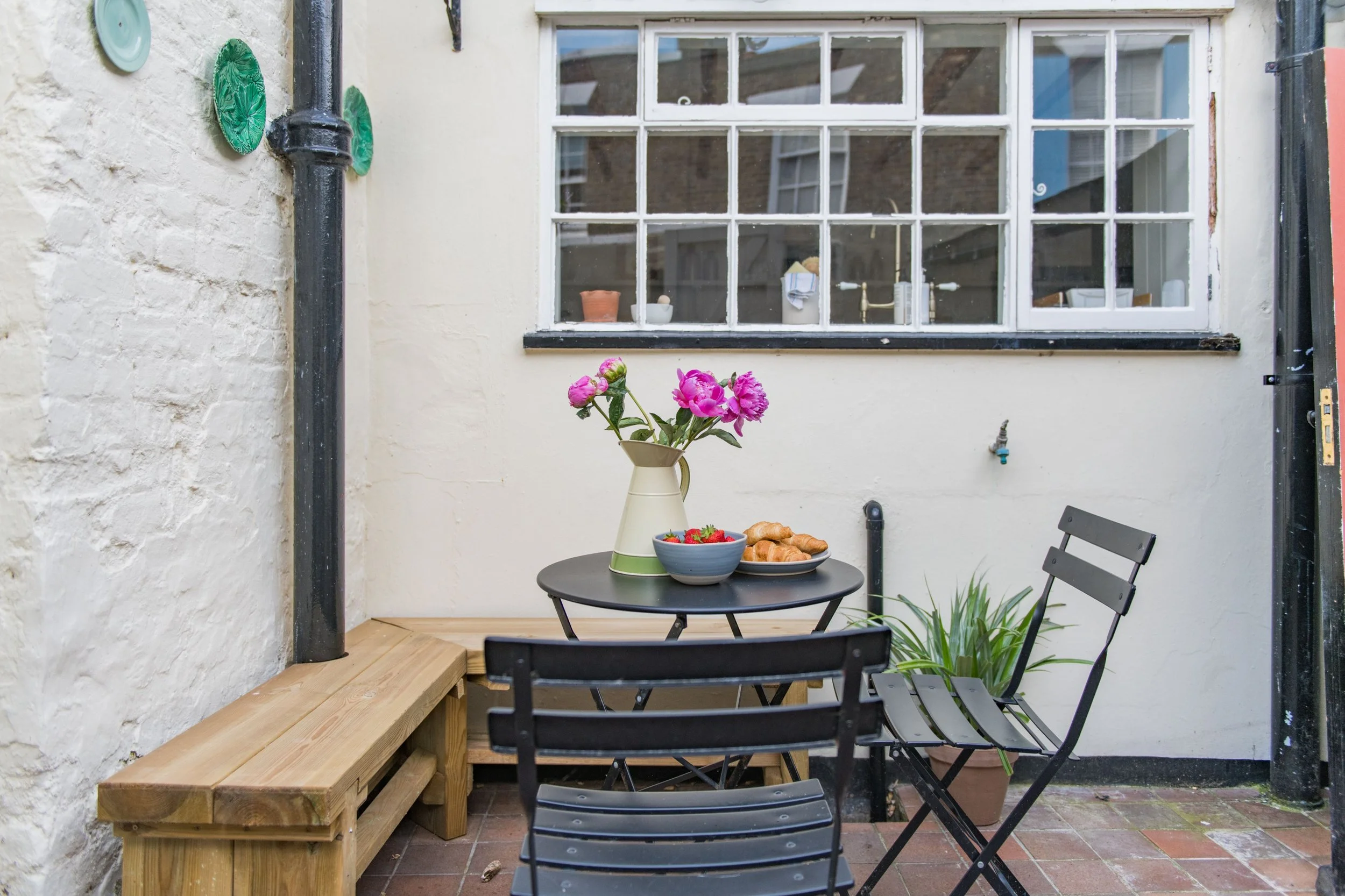 Small outdoor patio with a round table, two black metal chairs, and a wooden bench. On the table are a white pitcher with pink flowers, a blue bowl of strawberries, and a plate of croissants. There is a potted plant with long green leaves on the floo