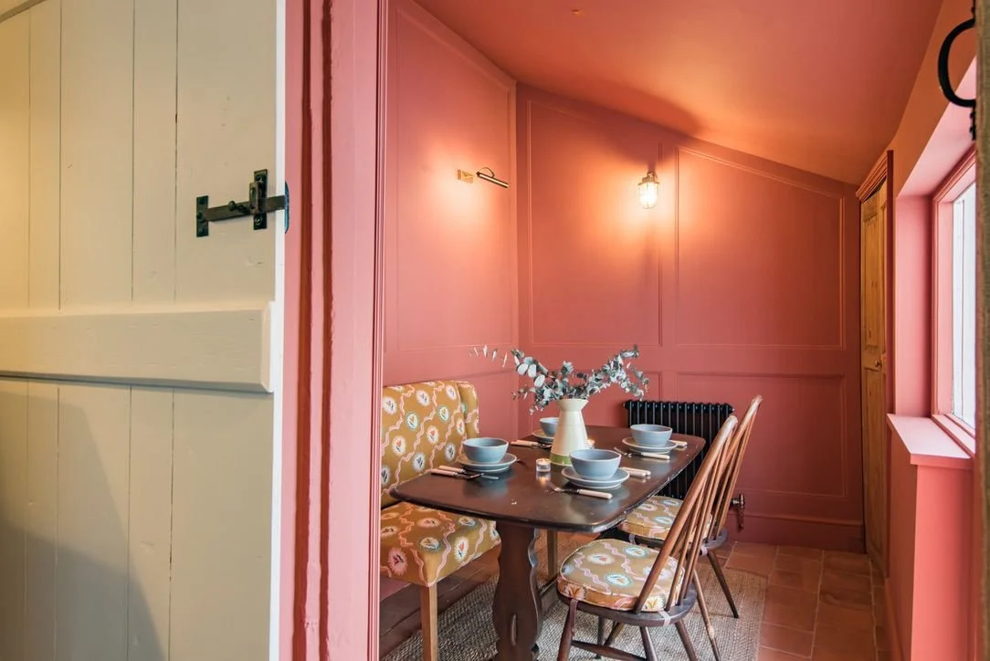 A cozy dining nook with pink paneled walls, a wooden table set with four blue dishes and cutlery, a yellow patterned bench, and three wooden chairs. A white vase with greenery is in the center of the table, and there is a window on the right side let