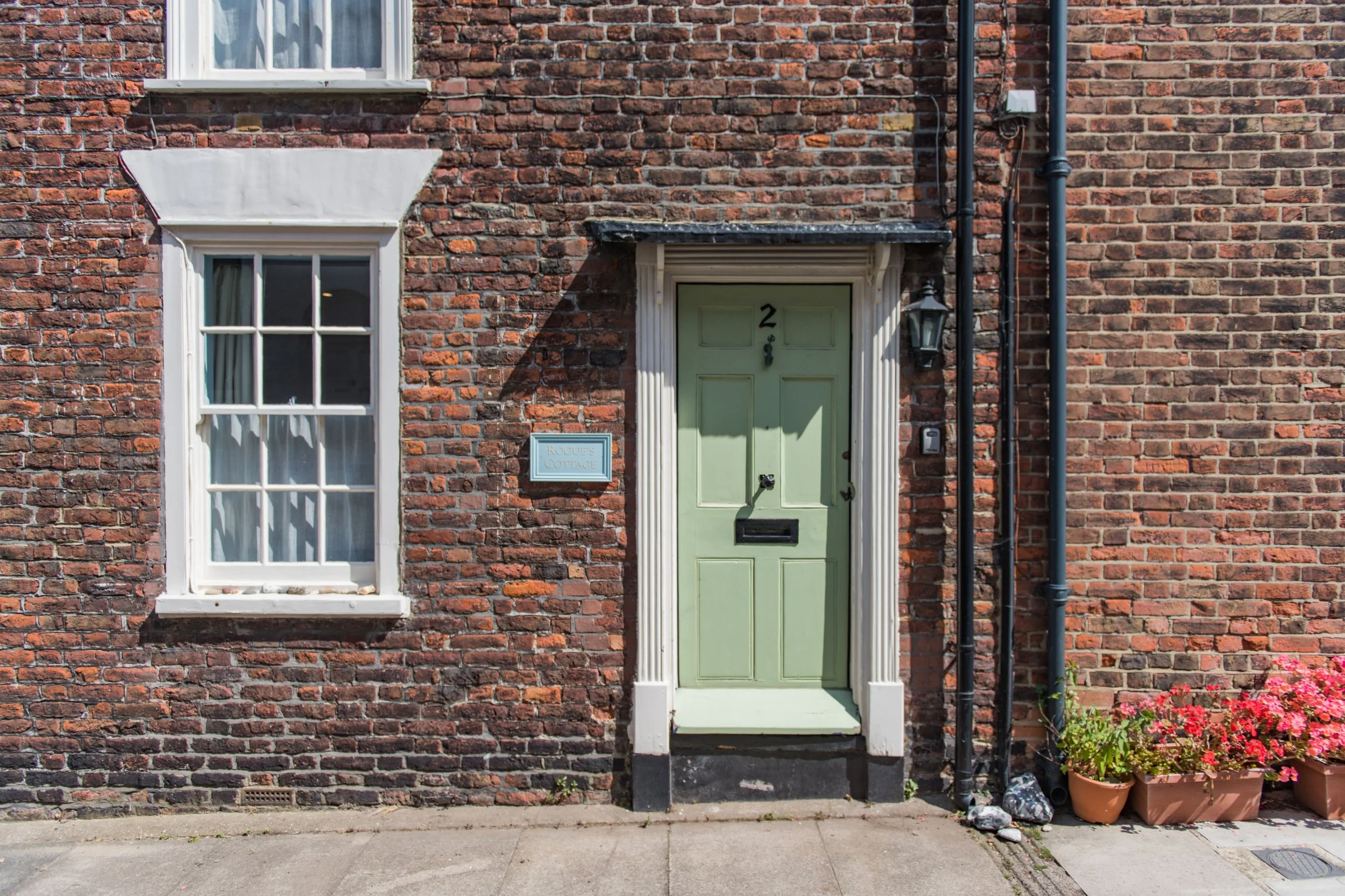 Brick house facade with green front door, white-framed window, potted flowers, and a sign that reads 'Rogue's Cottage'.