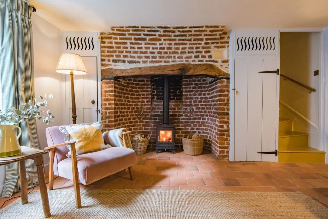 Living room with a brick fireplace, pink armchair, floor lamp, side table with green pitcher and flowers, curtains, and yellow staircase.
