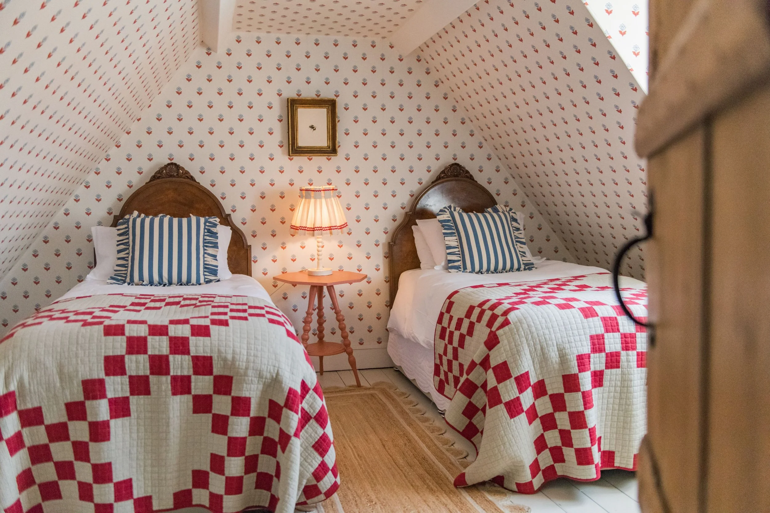 A cozy bedroom with two twin beds with wooden headboards, striped pillows, and quilted bedspreads featuring red and white checkered patterns, a small wooden nightstand with a table lamp, floral wallpaper, a small picture frame on the wall, and a beig
