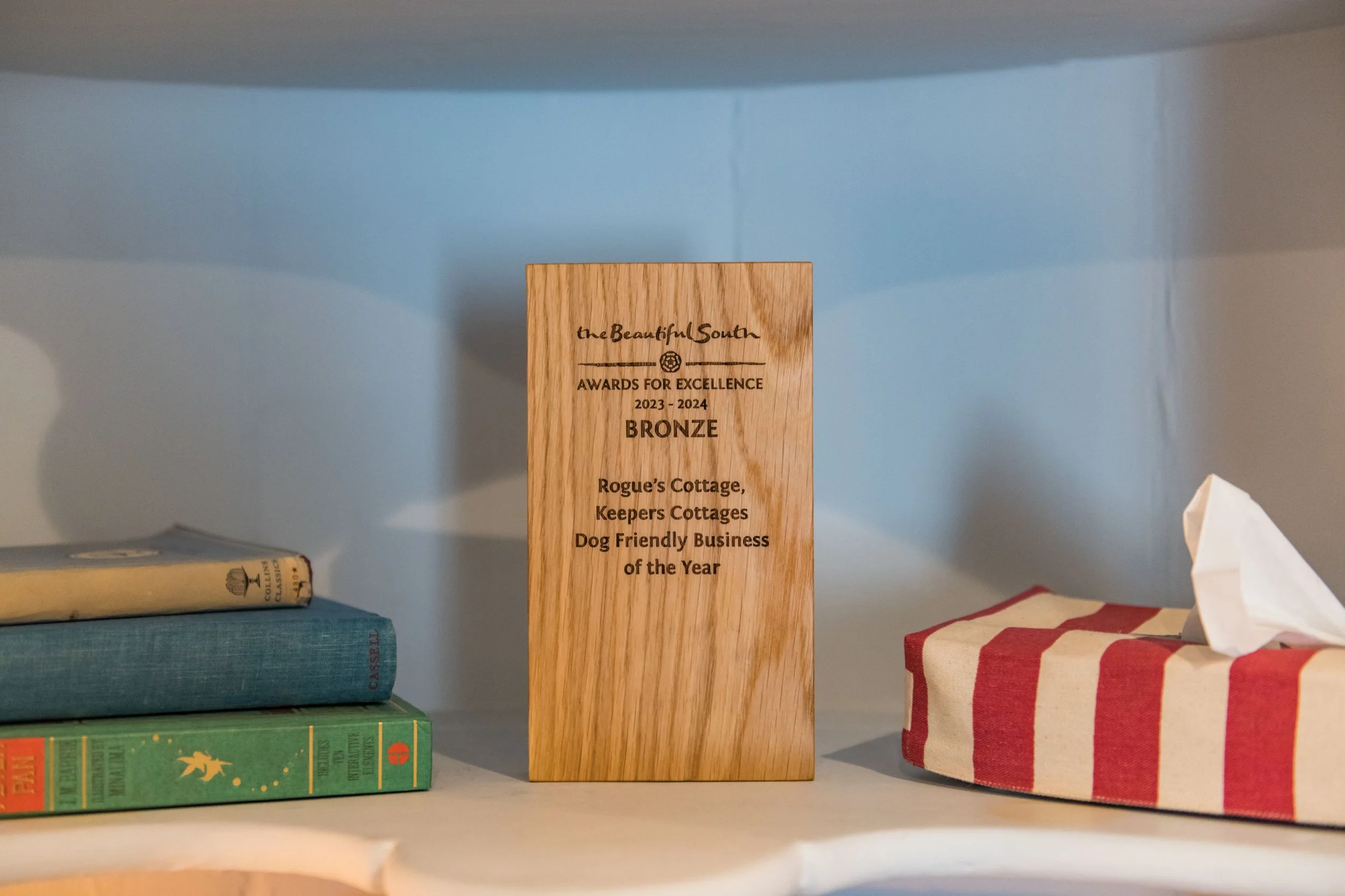 A wooden award plaque that reads 'the Beautiful South Awards for Excellence 2023-2024 Bronze, Rogue's Cottage, Keepers Cottages, Dog Friendly Business of the Year' placed on a white shelf. To the left, there are three books stacked on top of each oth