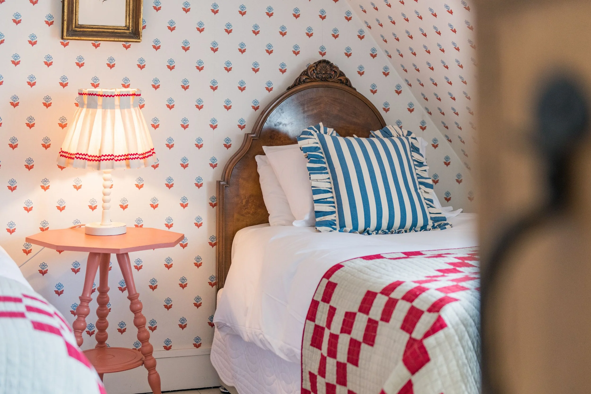 A bedroom with vintage-style floral wallpaper, a wooden headboard, a blue and white striped pillow with ruffled edges, a quilt with red and white checkered pattern, a pink side table, and a table lamp with a pleated shade.
