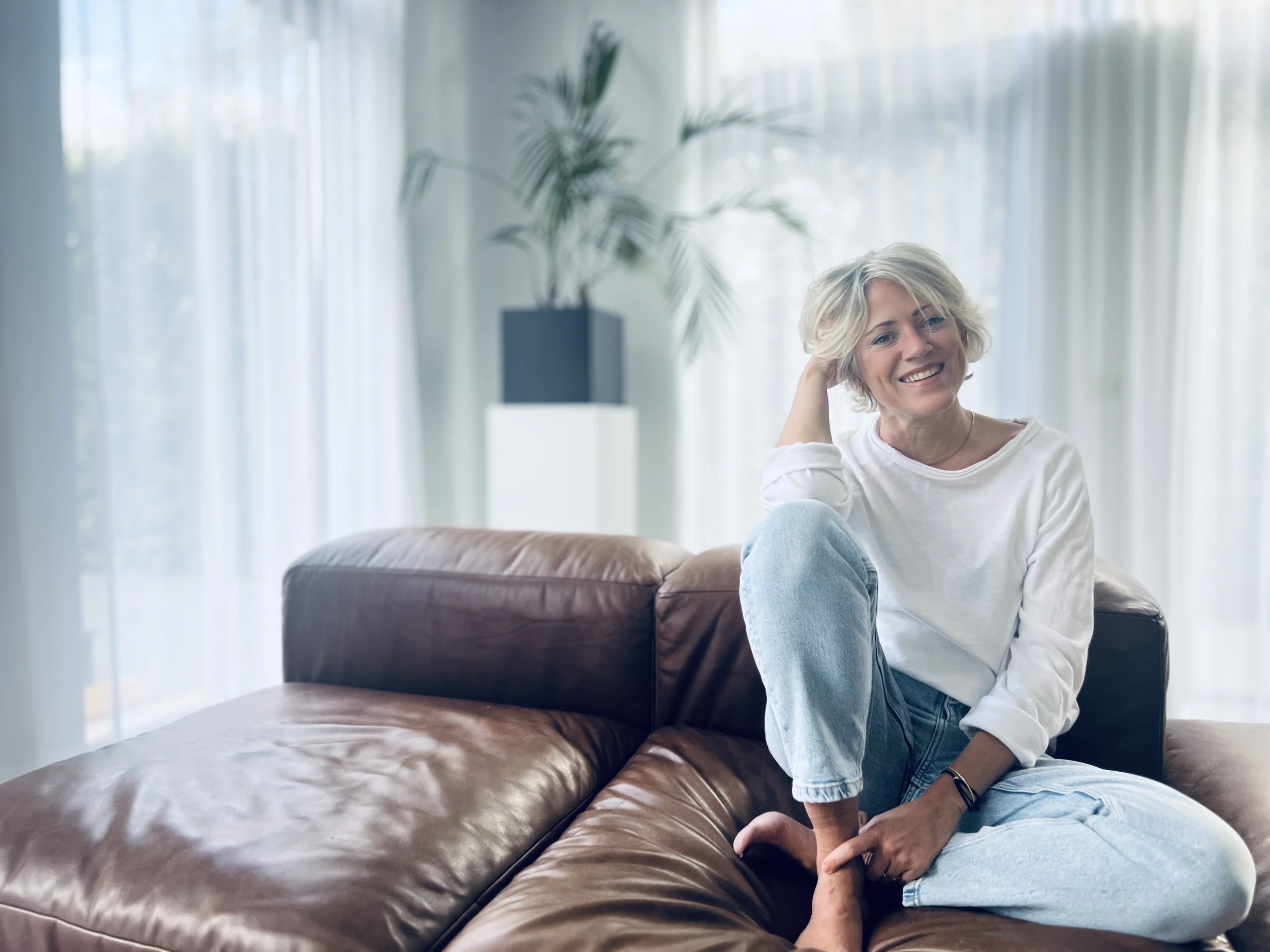 Florien, oprichtster van Ayurflo, zittend op een bank in een lichte woonkamer met grote ramen en planten, ontspannen en glimlachend.