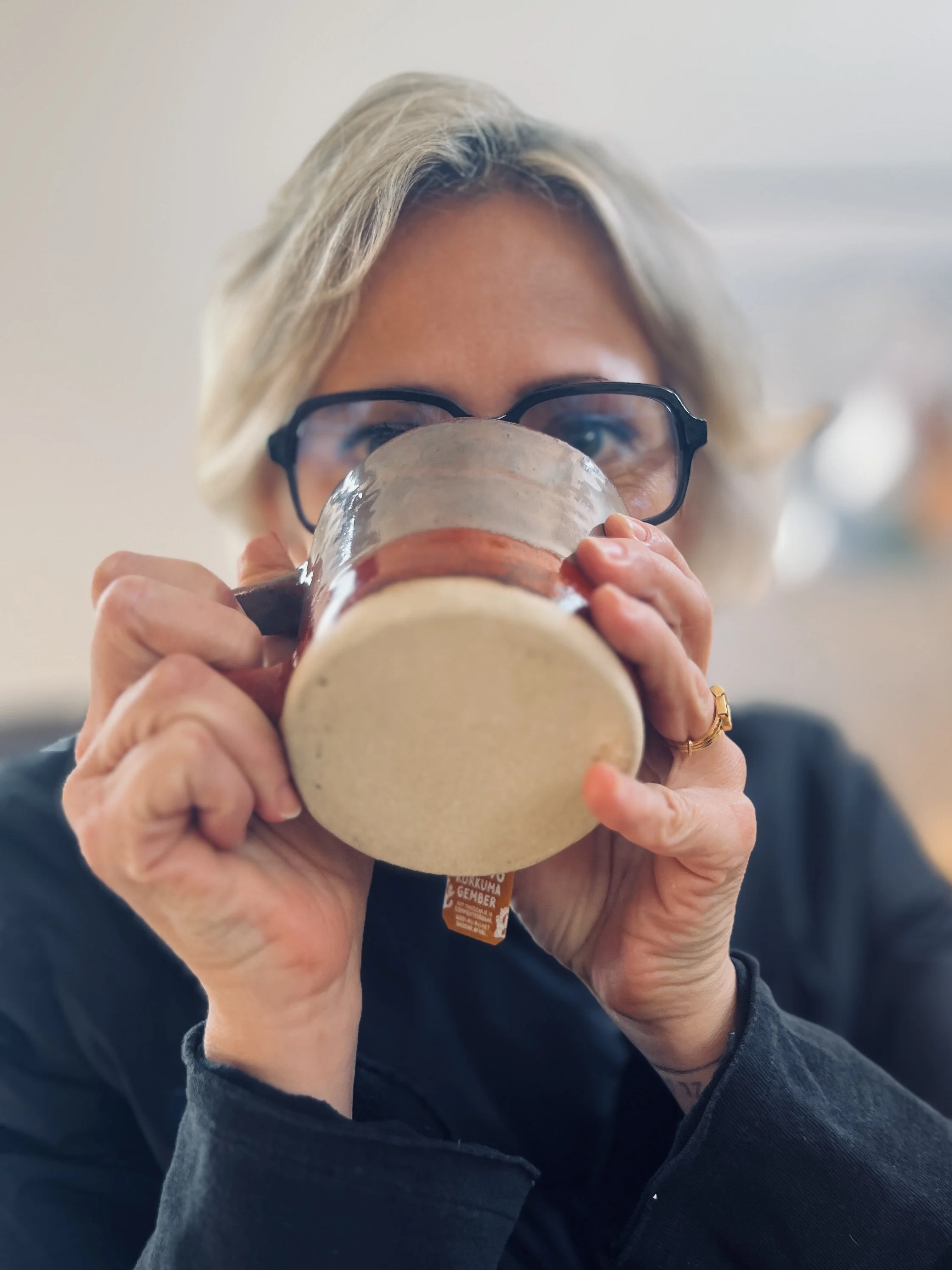 Vrouw die rustig een warme drank drinkt en in de camera kijkt, in een ontspannen setting.