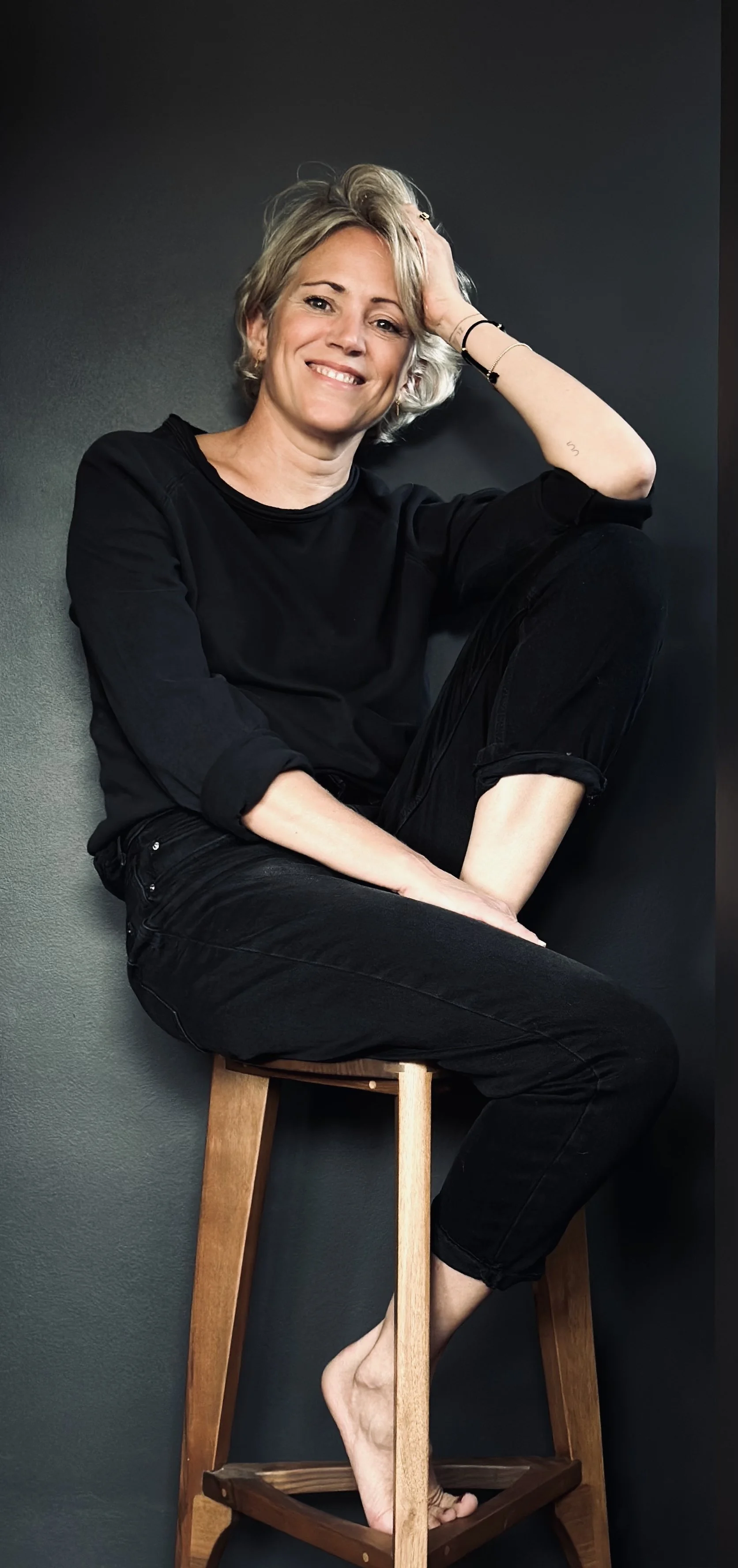 Florien, oprichtster van Ayurflo, zittend op een houten kruk tegen een donkere achtergrond, ontspannen en glimlachend.