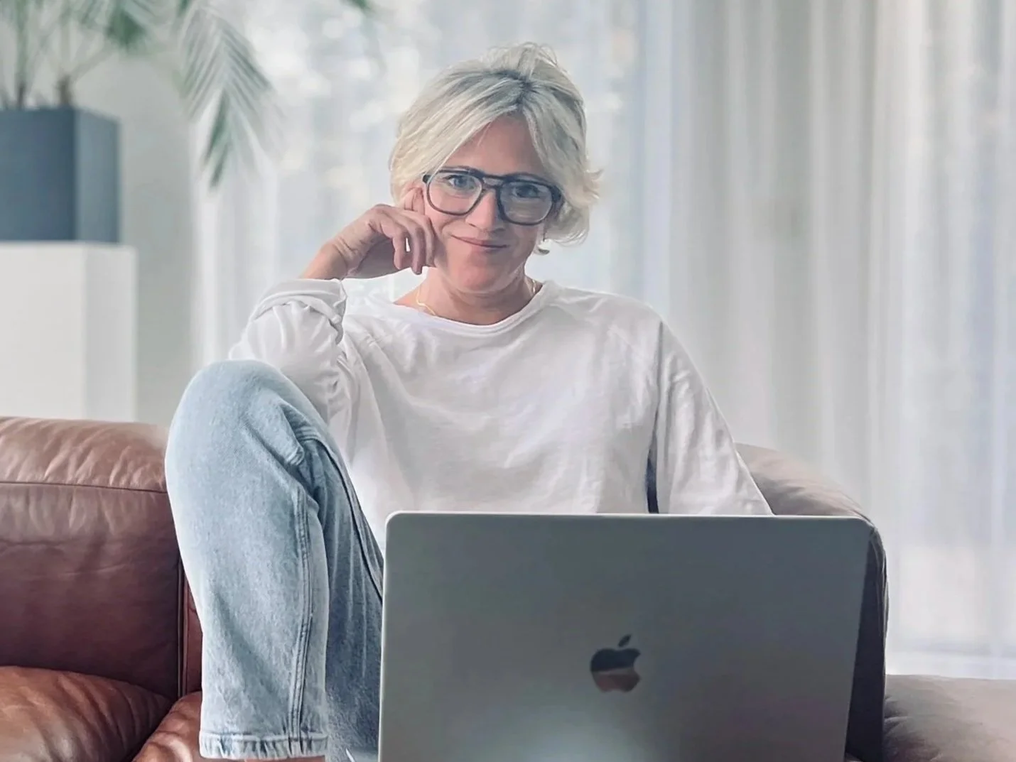 Florien, oprichtster van Ayurflo, zittend op een bank met een laptop in een lichte kamer met planten, ontspannen en geconcentreerd.