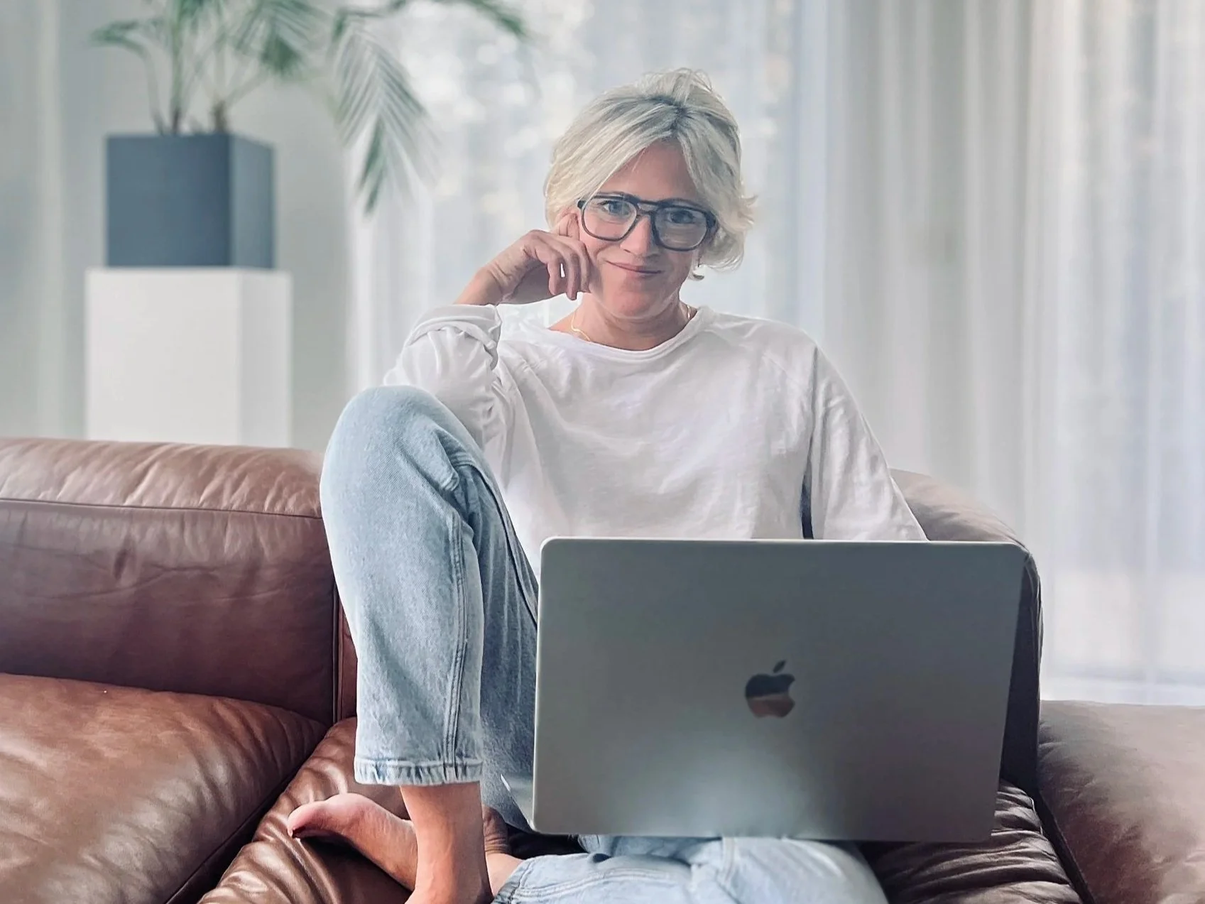Florien, oprichtster van Ayurflo, zittend op een bank met een laptop in een lichte ruimte met planten, in een rustige werkomgeving.