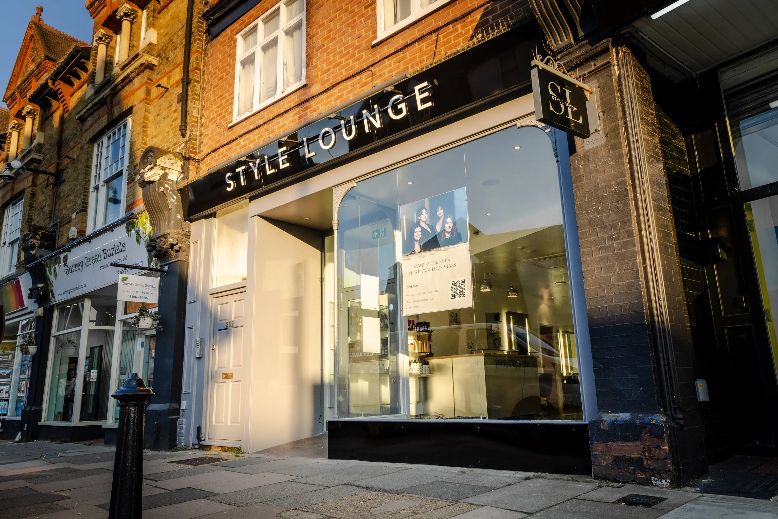 Storefront with a sign reading 'STYLE LOUNGE' above large glass windows reflecting the sky and nearby buildings, a black exterior with decorative column details, and part of a neighboring store with a white sign and merchandise visible inside.