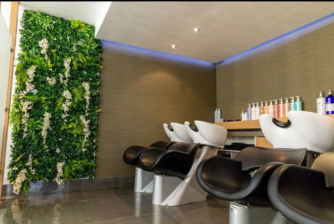 Salon with black and white shampoo bowls and chairs, a green wall with plants and white flowers, and bottles of hair products on a wood countertop.