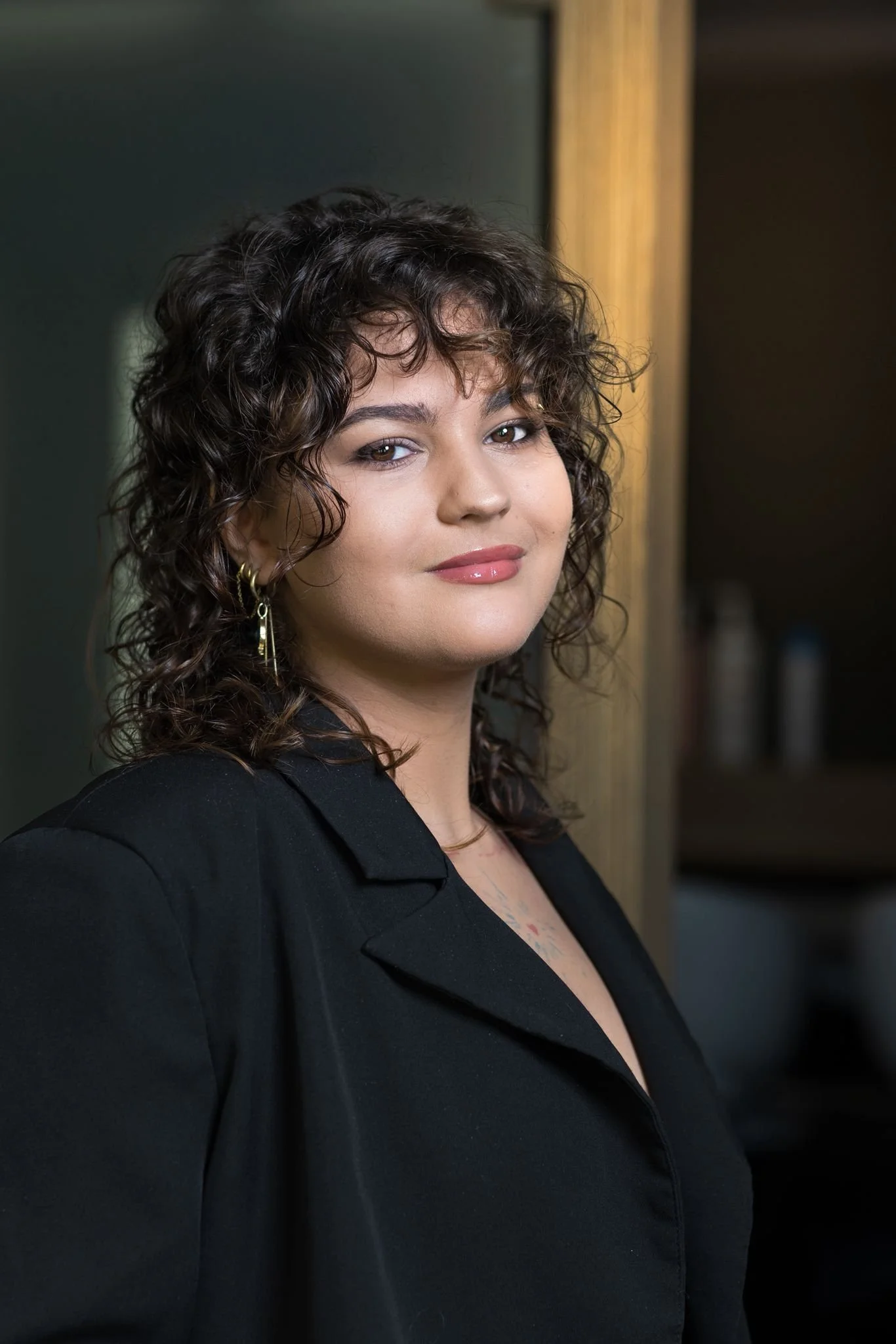 A woman with shoulder-length curly brown hair, wearing a black top, earrings, and makeup, standing indoors near a wooden cabinet.