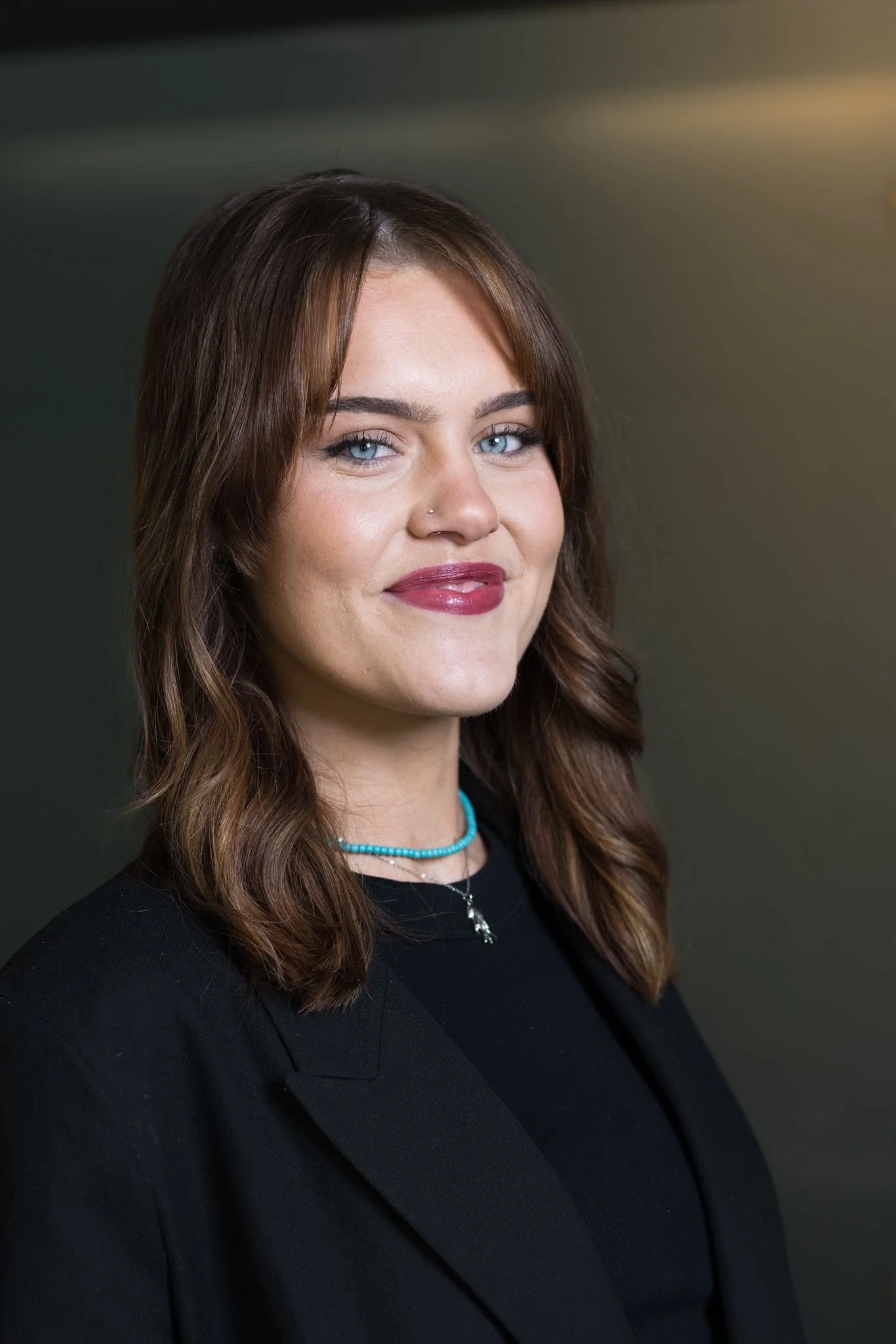 A woman with shoulder-length brown hair and blue eyes, wearing a black blazer, turquoise necklace, and a silver necklace with a cross pendant, smiling against a dark gradient background.
