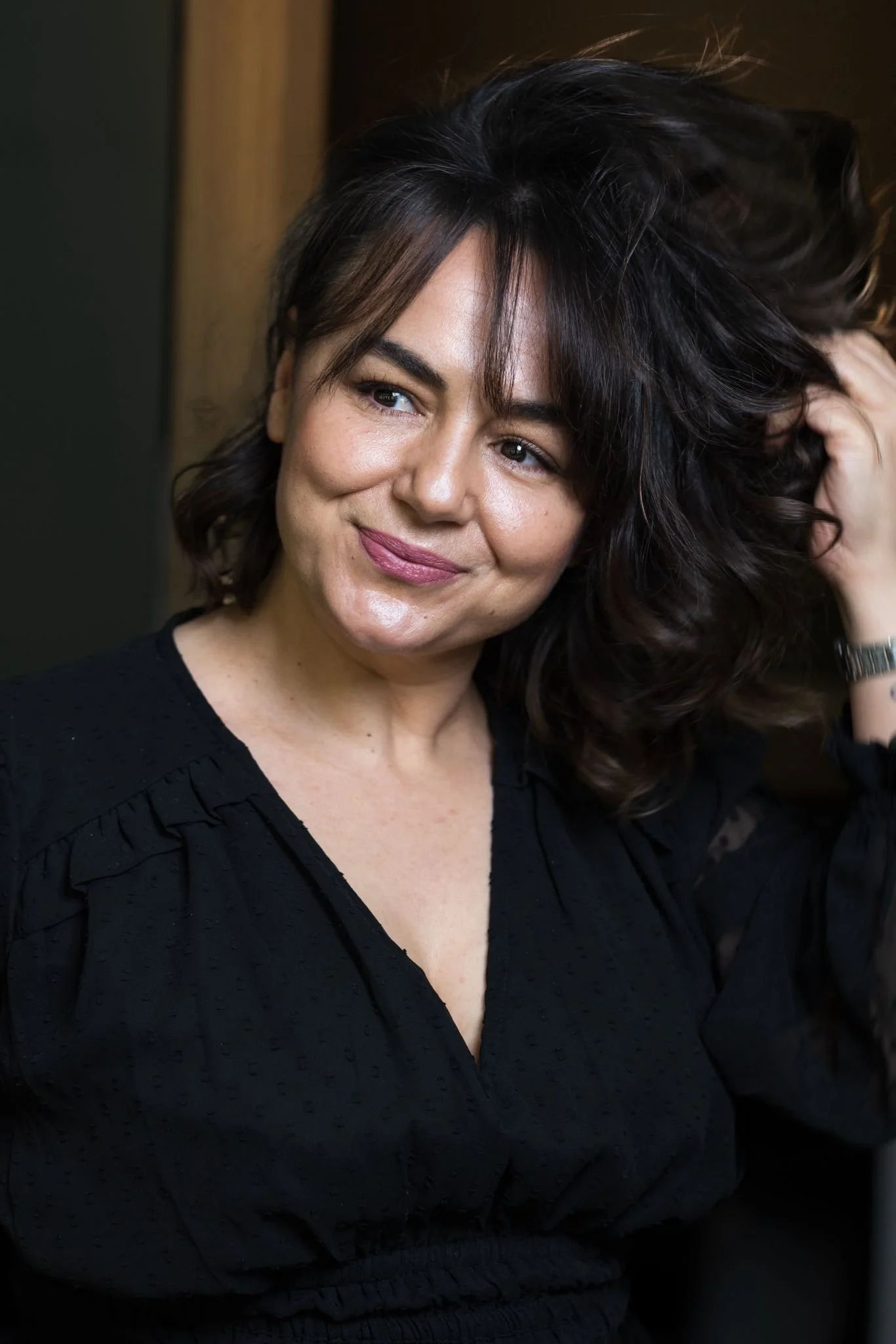 A woman with dark wavy hair and light skin, wearing a black blouse, smiling and touching her hair.