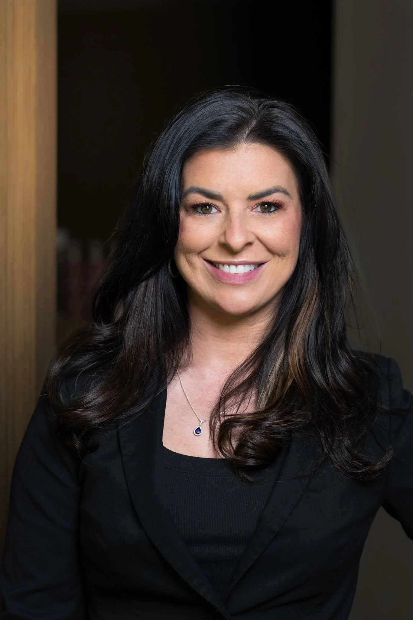 A woman with long dark hair, smiling, wearing a black blazer and a necklace with a dark blue pendant, standing indoors.