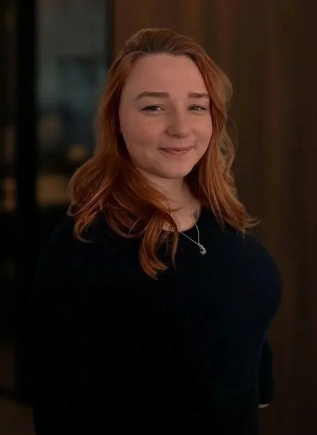 A woman with light skin and long reddish hair smiling softly, wearing a black top and a necklace, standing indoors with a blurred background.