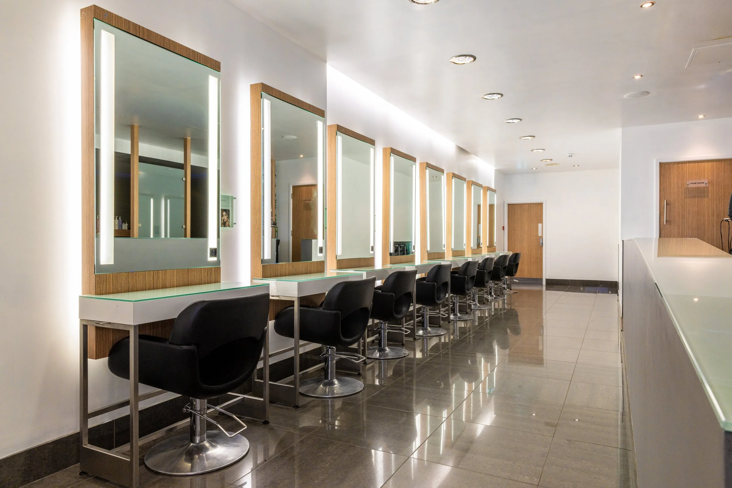 A row of black styling chairs in front of mirrors with wooden borders and built-in lights, located in a modern salon or beauty studio.