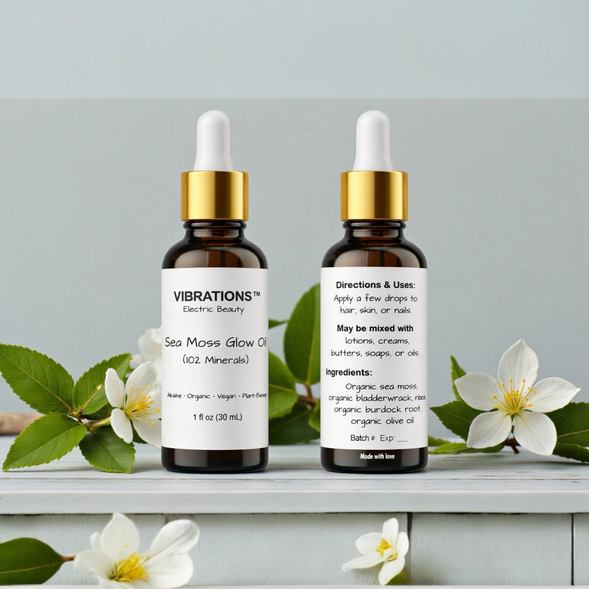 Two small brown bottles with white labels and gold caps, one showing the front label and the other showing the back label, surrounded by white flowers and green leaves on a white wooden surface.