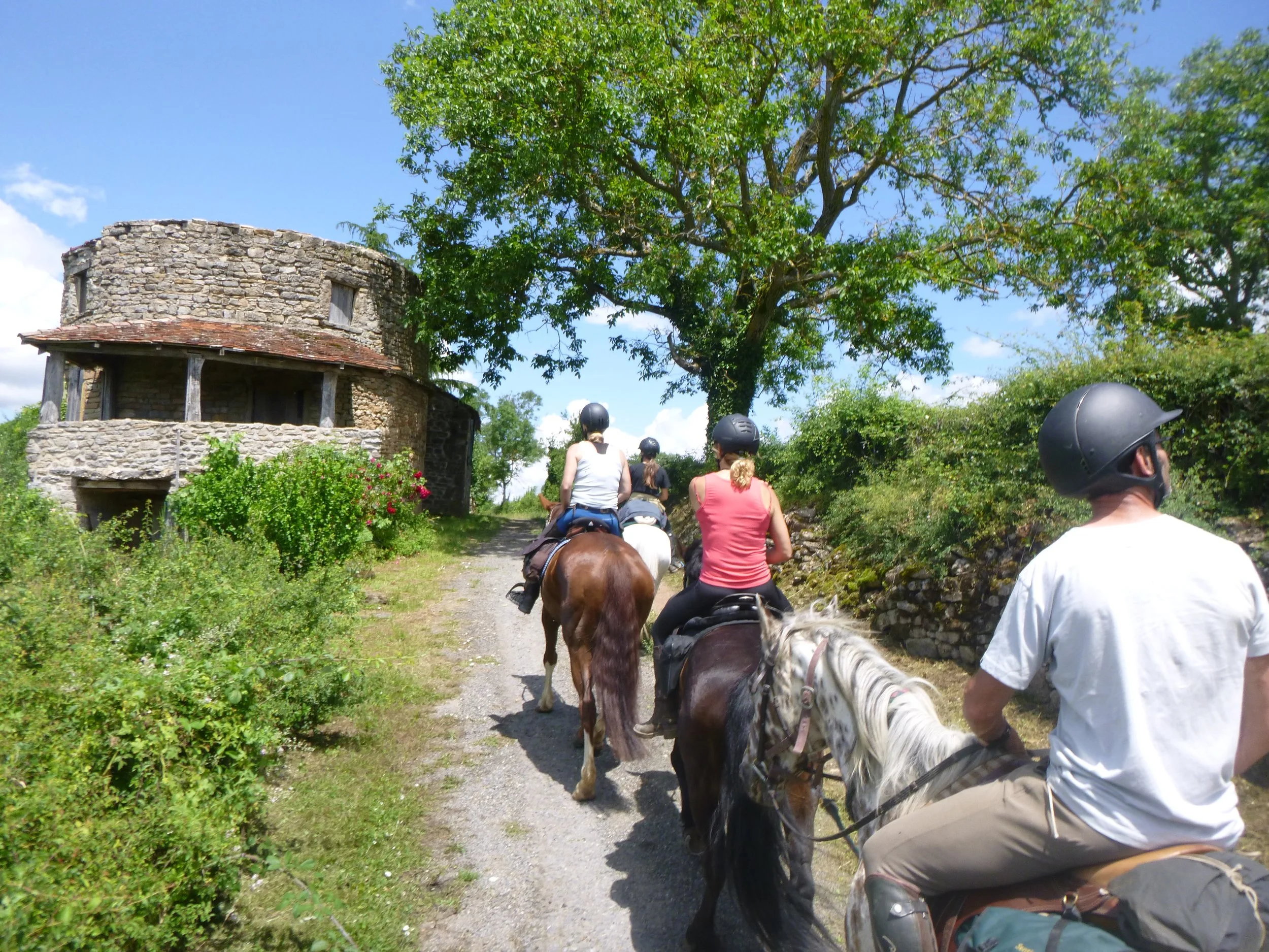 Groupe de personnes faisant une balade à cheval sur un sentier bordé d'arbres et de buissons, avec une vieille maison en pierre en arrière-plan.