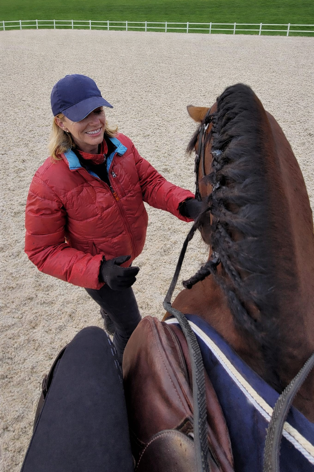 Une femme souriante en veste rouge et casquette bleue, en train de caresser un cheval marron avec une longue crinière tressée, dans une écurie avec clôtures blanches.