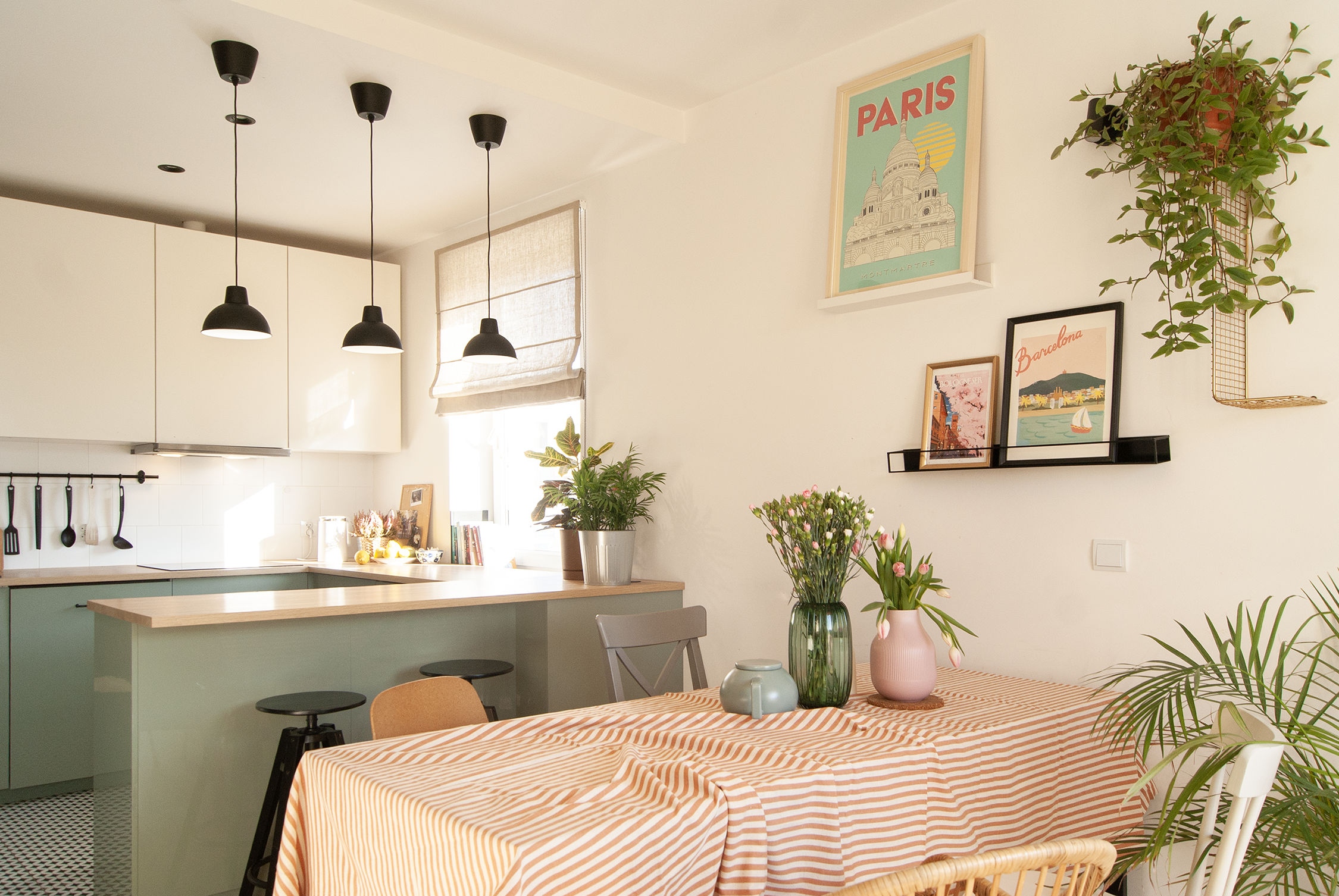 A cozy kitchen and dining area with a table covered in an orange and white striped tablecloth. On the table are vases with flowers and small decorative items. The background features a kitchen with white cabinets, black pendant lights, and a window with a beige roman shade. Decor on the wall includes framed pictures and a large poster of Paris, with hanging plants adding greenery to the space.