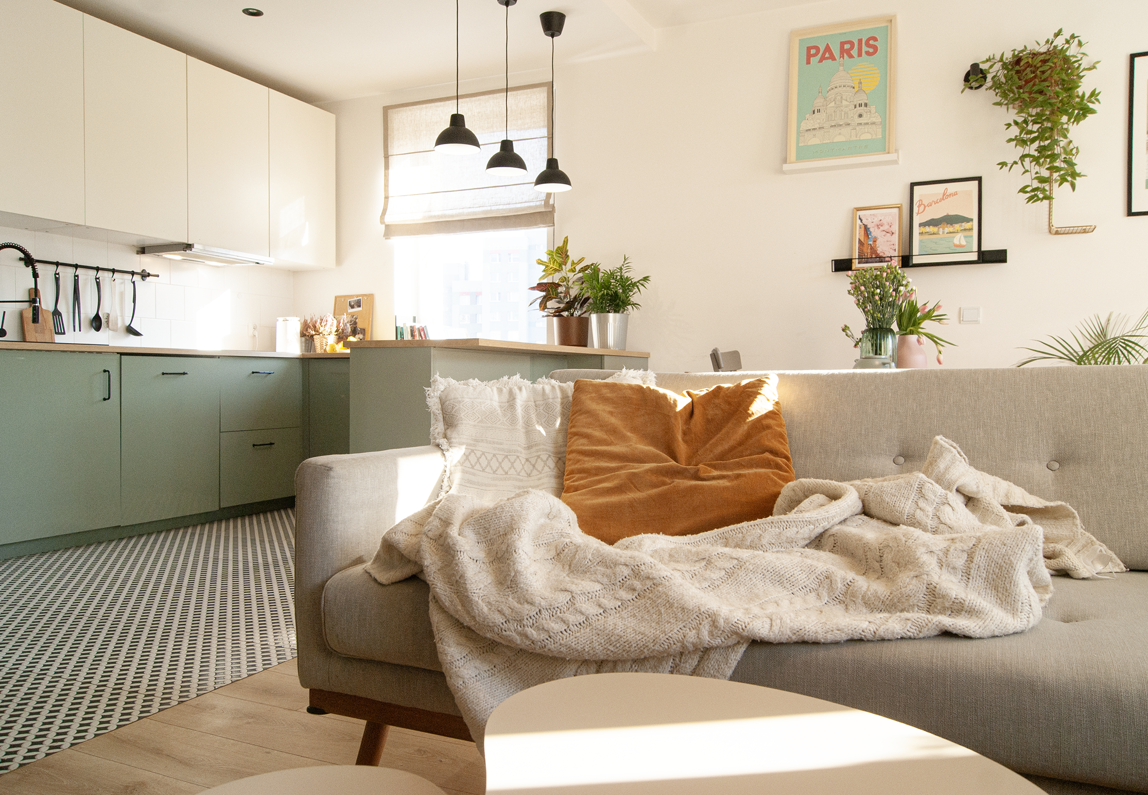 Cozy living room with a beige sofa, orange and cream pillows, a knitted blanket, open kitchen with green cabinets, and decorative wall art featuring Paris and Barcelona posters.