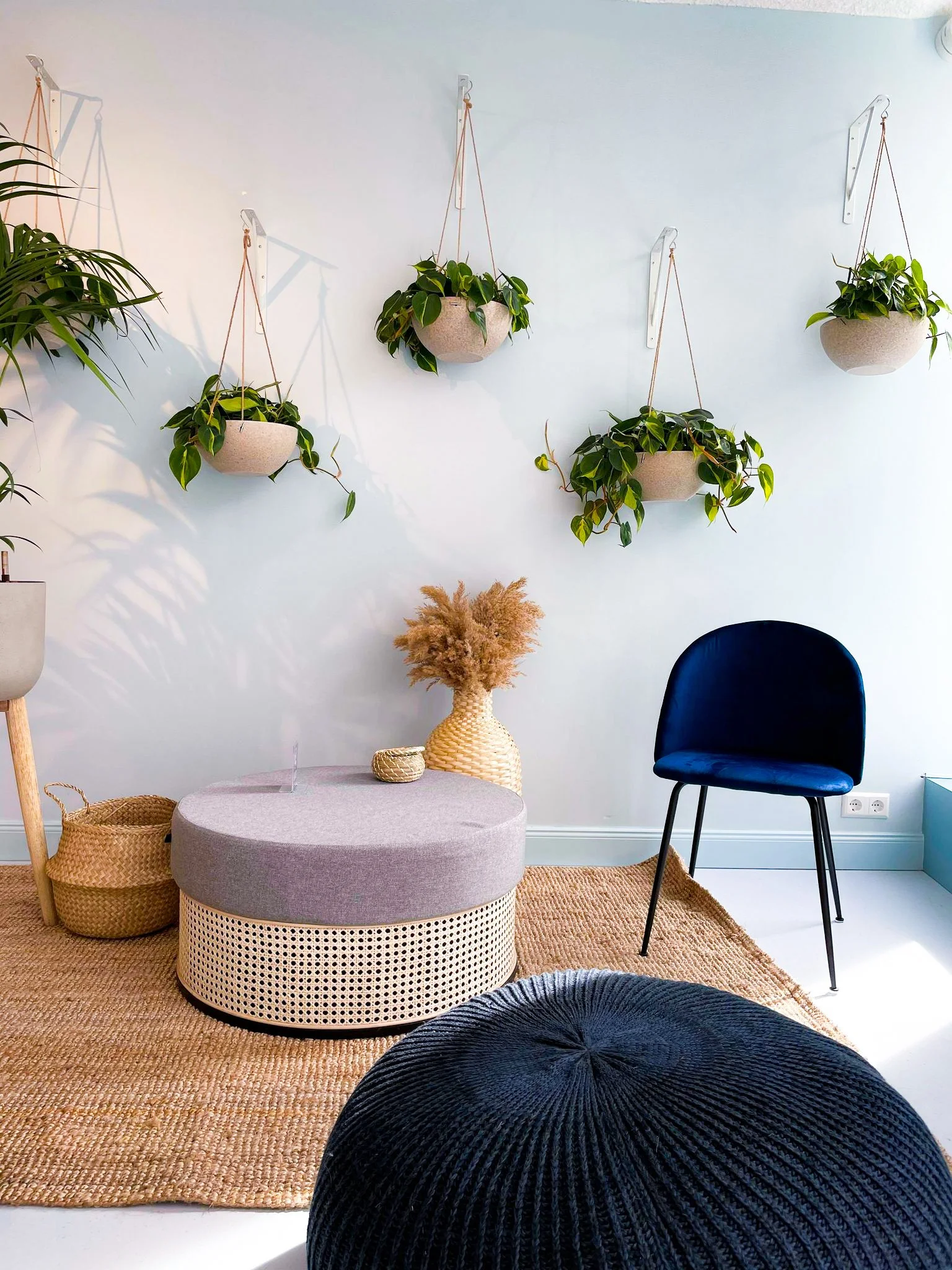 Interior living space with hanging potted plants on the wall, a black velvet chair, a round beige ottoman with a gray cushion, a wicker vase with pampas grass, a woven basket, and a black pouf on a jute rug.