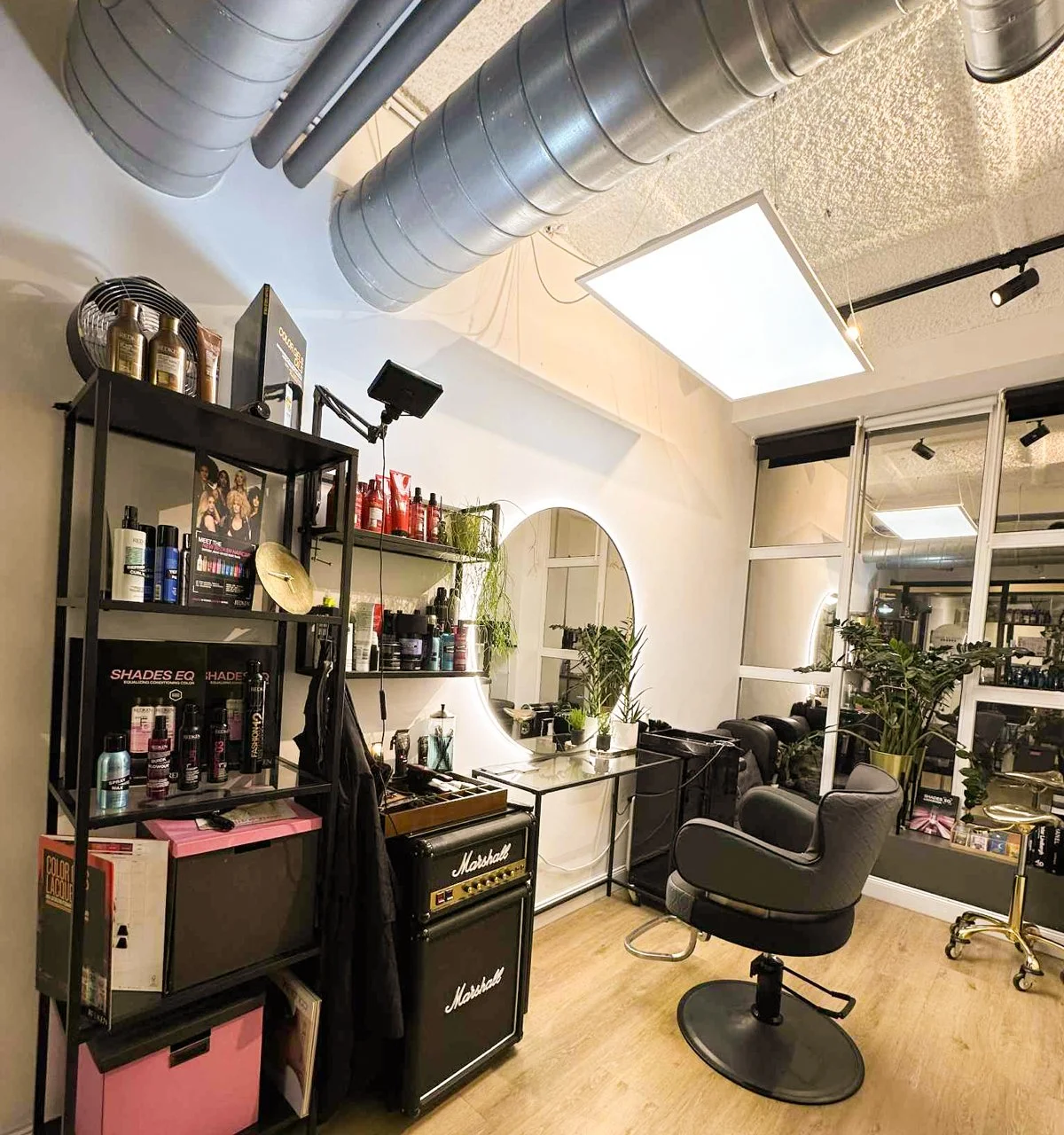 A hair salon with a styling chair, shelves with hair products, a mirror, plants, a Marshall amplifier, and large vents on the ceiling.